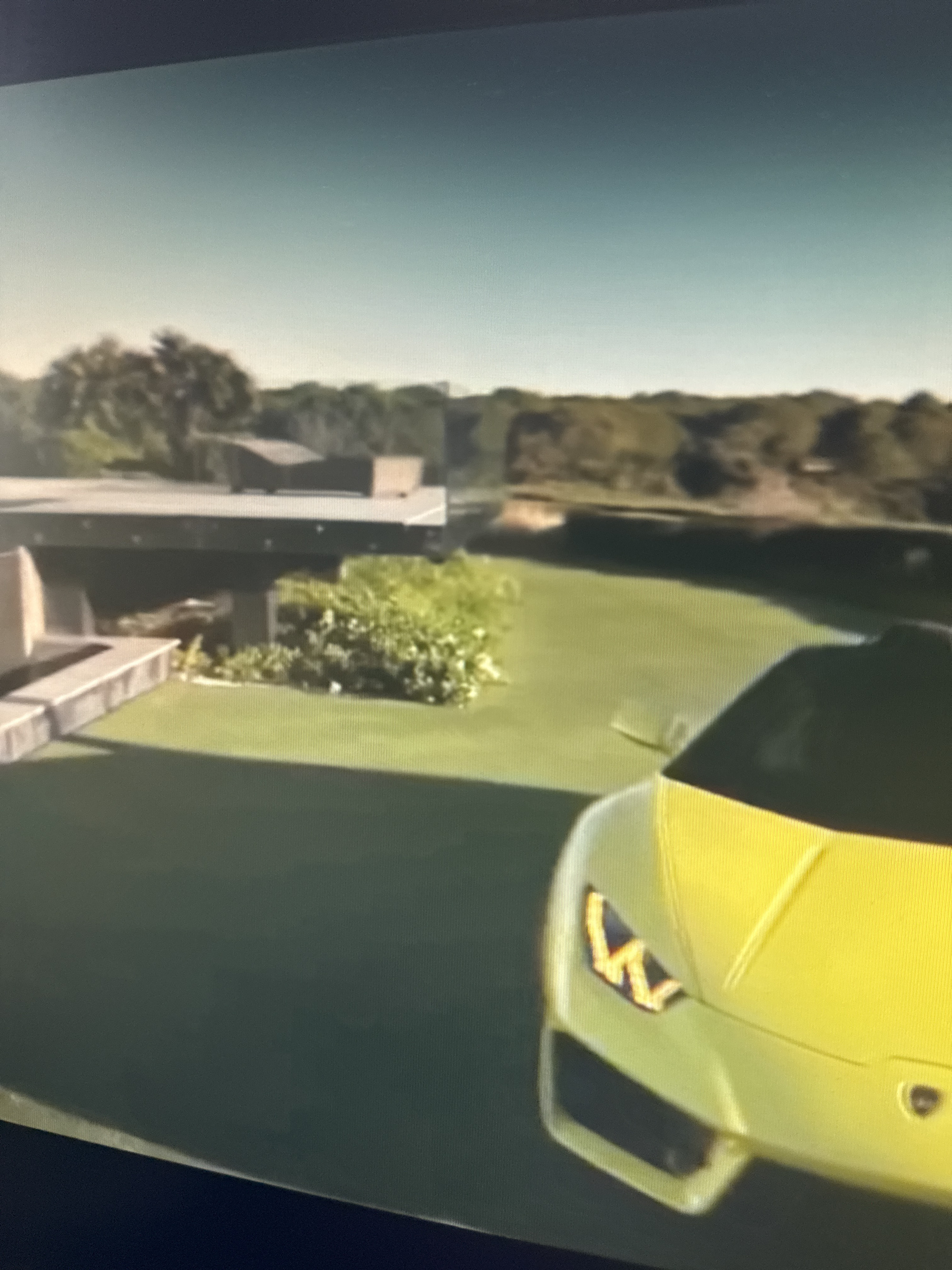 An aerial, slightly angled, medium-shot captures a vibrant lime-green sports car, likely a Lamborghini, parked on a neatly manicured green lawn. The car's front is partially visible, revealing its distinctive headlights and sleek design. Behind the car, a modern building with flat, gray roofing and concrete accents stands on the left side of the frame. Lush green foliage and trees surround the building and extend into the background, forming a dense, wooded landscape under a hazy, pale blue sky. The overall scene suggests a luxurious property, possibly a private residence or a high-end establishment, situated in an upscale area with abundant greenery. However, without further context or identifiable landmarks, pinpointing a specific location within Netanya, Israel, is not possible based solely on this image.