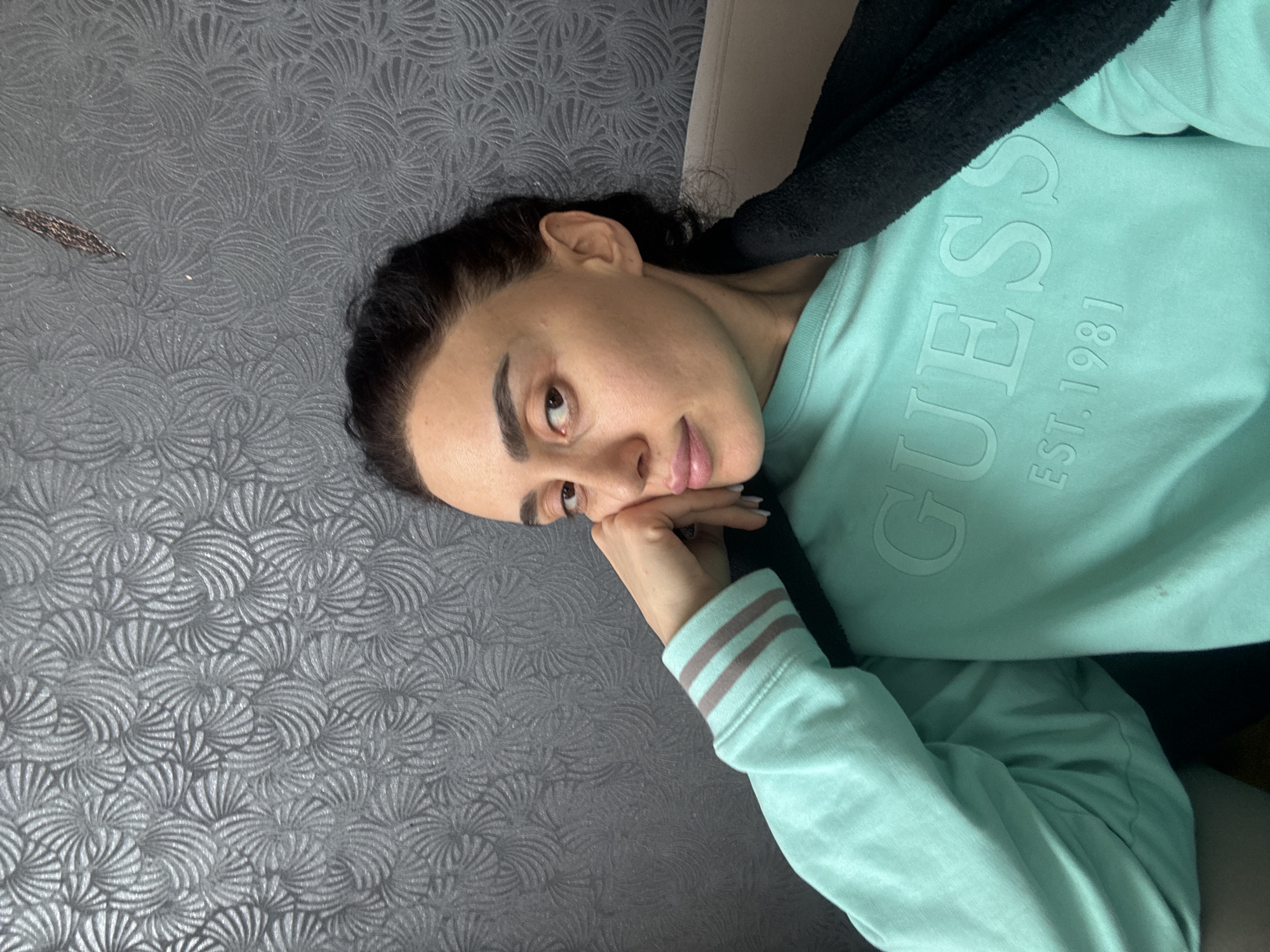 A woman is lying on a gray patterned surface. She is wearing a light blue Guess sweatshirt with the year "EST. 1981" printed on it. Her head is tilted, and her hand is resting on her face. The background features a wall or surface with a swirling, textured pattern. A black hoodie is behind her neck. There is no indication of the location being Tbilisi, Georgia, in the image.