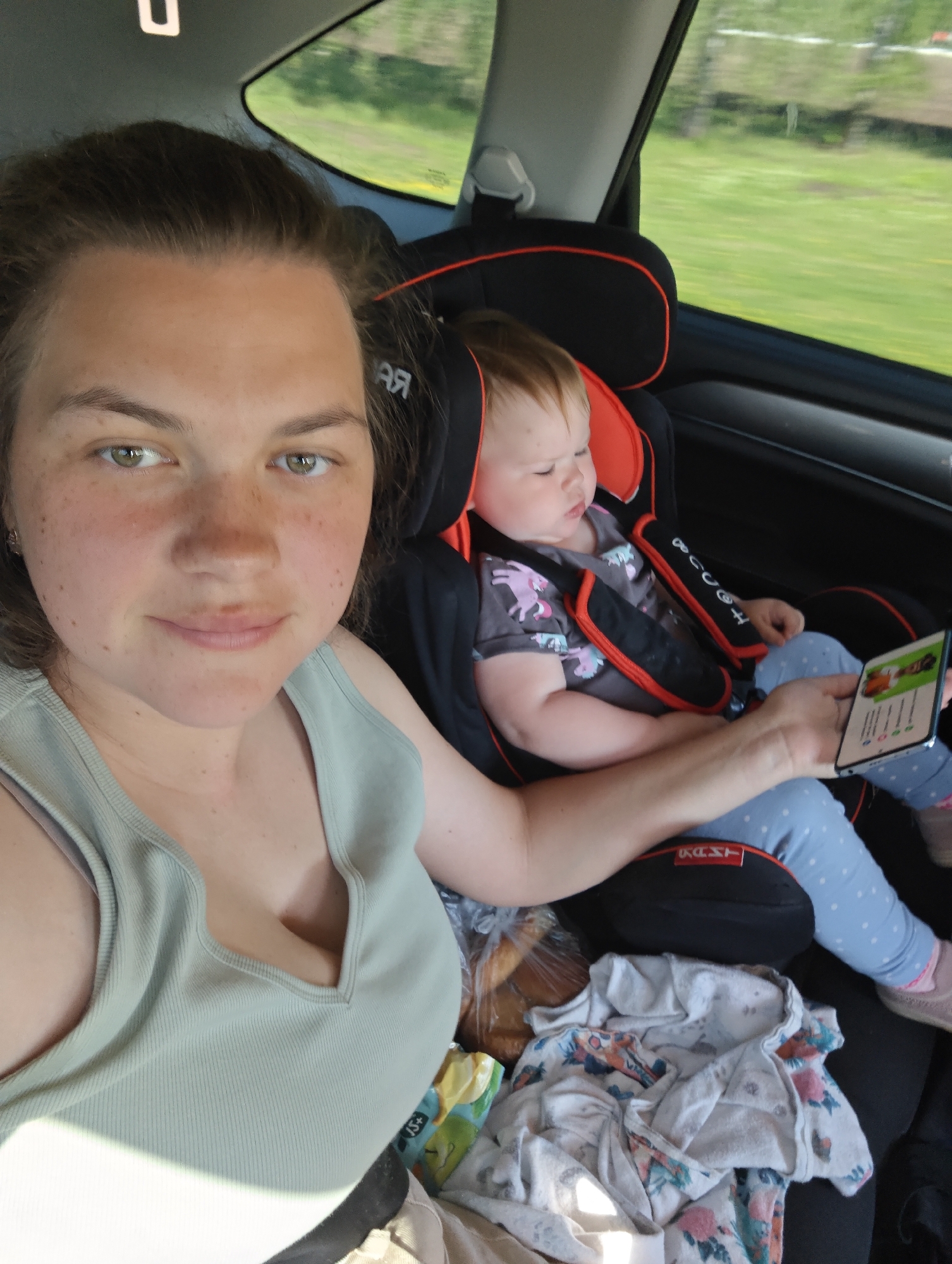 A woman and a young child are seated in a car. The woman is in the front seat, and the child is in a car seat in the back. The woman is looking at the camera, and the child is looking at a phone that the woman is holding. The car appears to be moving, and the background is blurred. The location is Agryz, Russia, as determined by EXIF data. The child is wearing a gray shirt and blue pants. There is a white floral jacket and a bag of bread on the seat next to the child.