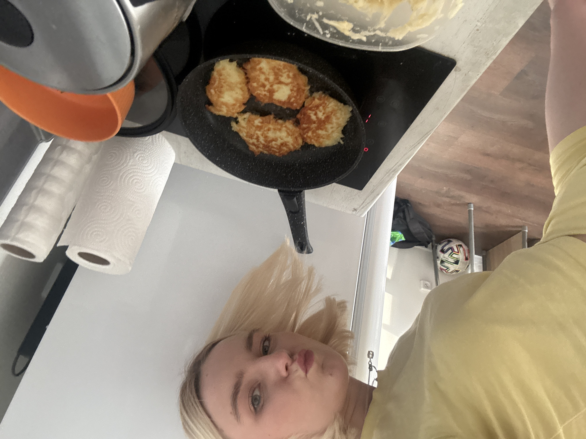 A young, fair-skinned person with blonde hair is facing the camera, wearing a yellow shirt. The camera's point of view is angled from above and to the side. The person is in a kitchen. Above and slightly behind them is a black frying pan on a black stovetop. Inside the pan are four golden-brown fried potato pancakes (latkes). A bowl of batter is partially visible above and to the right of the pan. To the left are a silver kettle with an orange spout and two rolls of paper towels. In the background, a soccer ball is visible on a shelf. The location is Unknown, Russia.