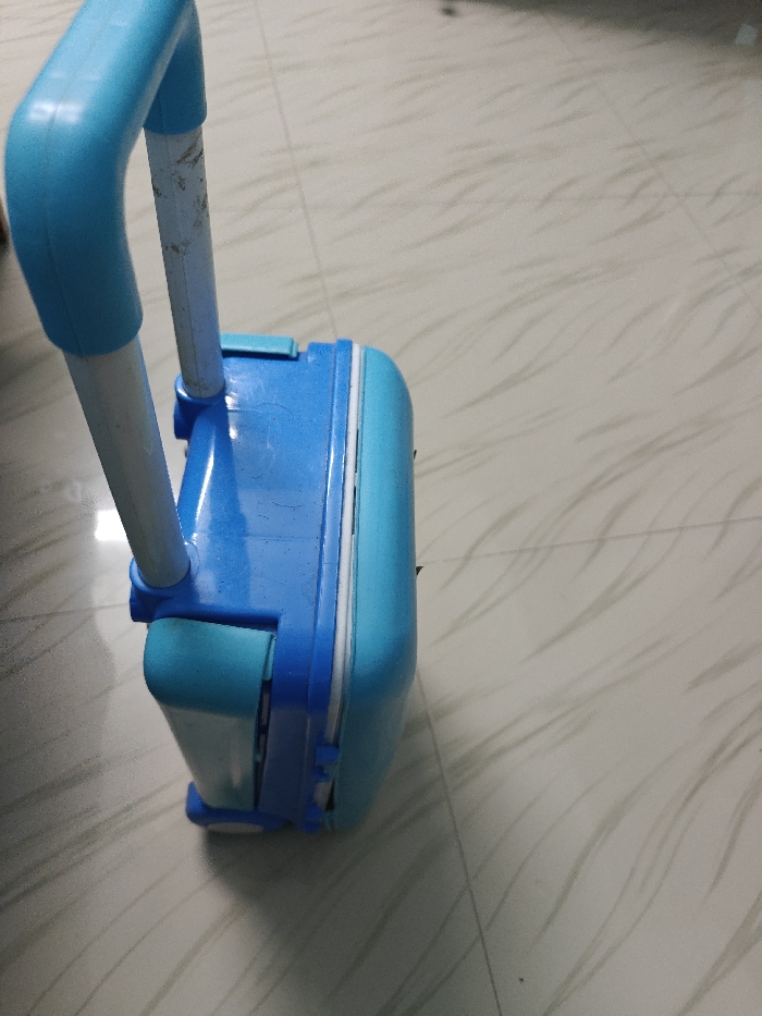 The image shows a blue and teal-colored suitcase with a white handle, lying on a tiled floor, positioned in a vertical orientation. The handle is extended and the suitcase is facing towards the right. The tiled floor has a white, grey, and beige pattern. The image was likely taken in a home, apartment, or hotel room in Tiruppur, India, based on the provided coordinates. There are no people or other objects in the image.