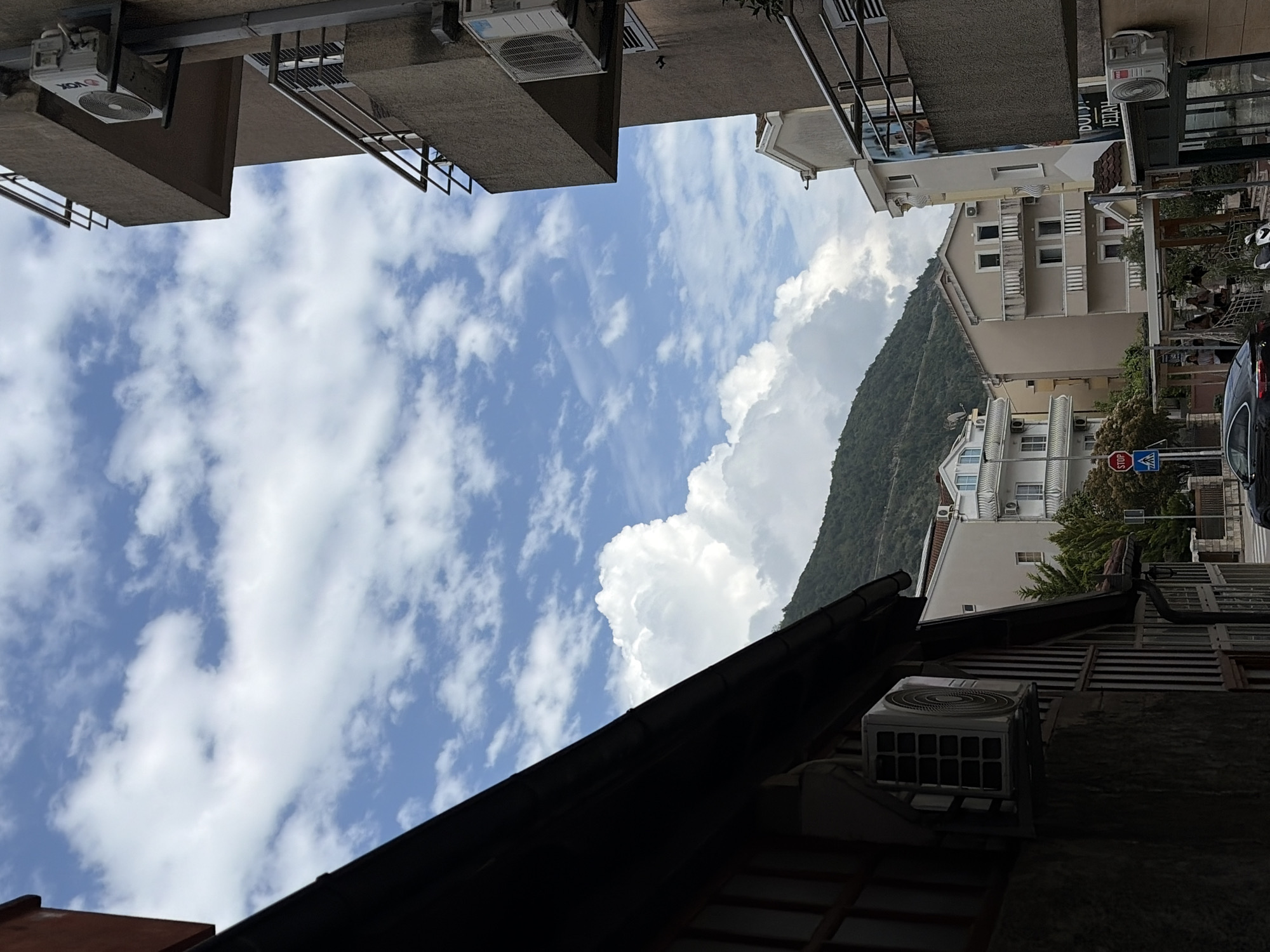 This daytime image captures a street scene in Budva, Montenegro, characterized by urban architecture set against a prominent mountainous backdrop. Buildings line both sides of what appears to be a street or pathway. On the left, multi-story structures with balconies, some casting deep shadows, are visible from a low angle. Several external air conditioning units are mounted on these facades, with one clearly displaying the "VOX" brand logo.
On the right side of the image, lighter-colored buildings with multiple windows and balconies are present, some appearing to have outdoor seating areas at street level. A large, green, tree-covered mountain dominates the background, stretching across the middle and right portions of the frame. Above the buildings and mountain, the sky is a bright blue, filled with numerous white, fluffy cumulus clouds, indicating fair weather.
Notable street elements include a red octagonal "STOP" sign and a blue pedestrian crossing sign visible on the right, near a partially obscured parked black car. The foreground features the dark, tiled roof of a building, partially framing the bottom of the scene. Faint, indistinct text "DULJI... JEDAN" is visible on a sign attached to one of the buildings. The sunlight creates clear shadows, suggesting the time of day is likely mid-morning to late afternoon. The scene is static, depicting the architectural and natural environment of the location.