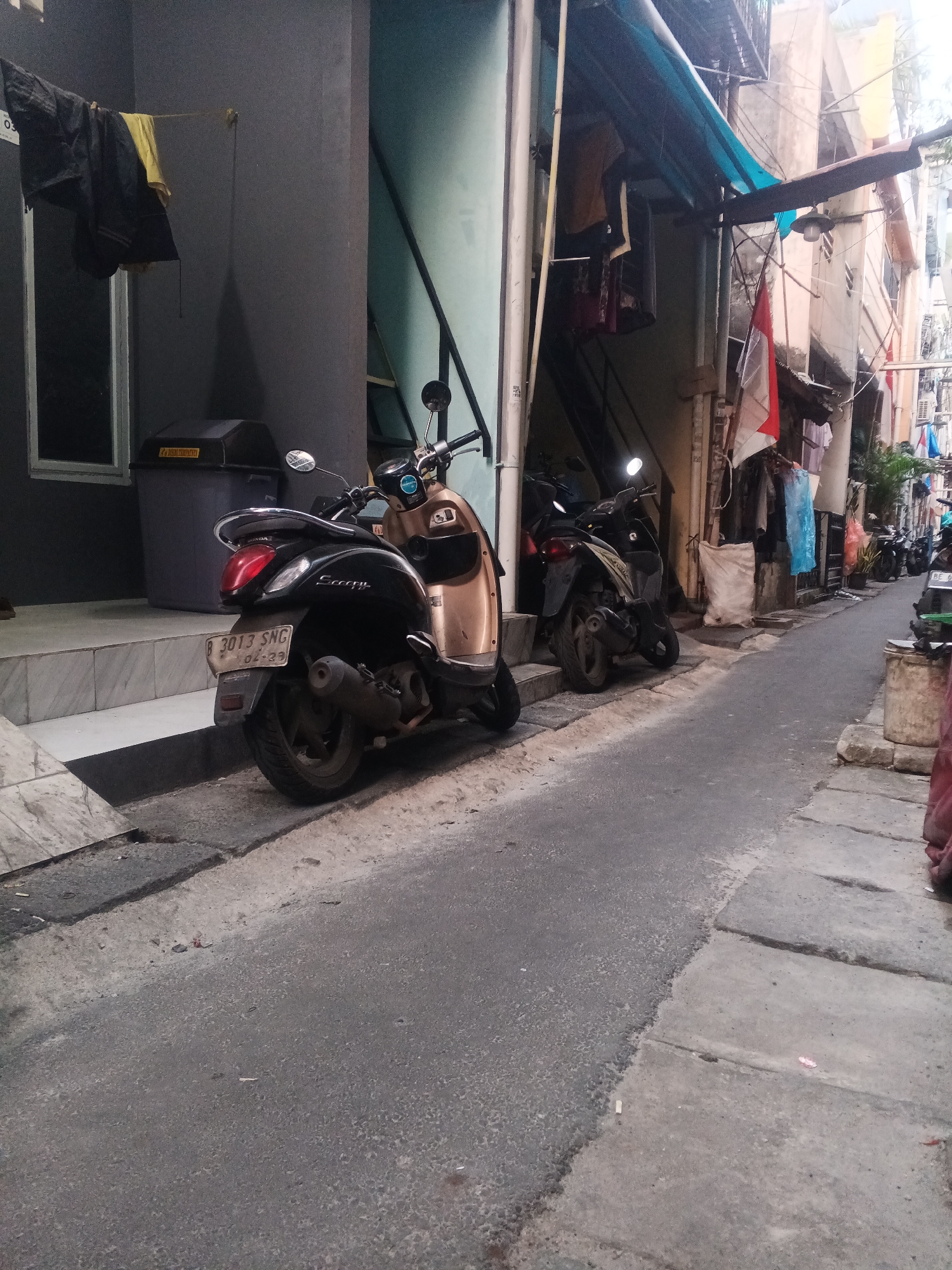 The image depicts a narrow alleyway in West Jakarta, Indonesia, lined with buildings and parked motorcycles. In the foreground, a black and gold scooter is parked on the side of the asphalt road, its license plate visible and reading "B 3013 SNG". To the left, a dark gray wall with a window and a trash bin is visible, with laundry hanging on a line in front of it. The alleyway is covered by awnings and wires overhead, with more motorcycles and buildings extending into the background. Clothes are hanging from various windows and lines, adding to the lived-in feel of the urban environment. The scene is illuminated by natural light, with the overall impression being that of a typical residential or commercial alley in a densely populated urban area.