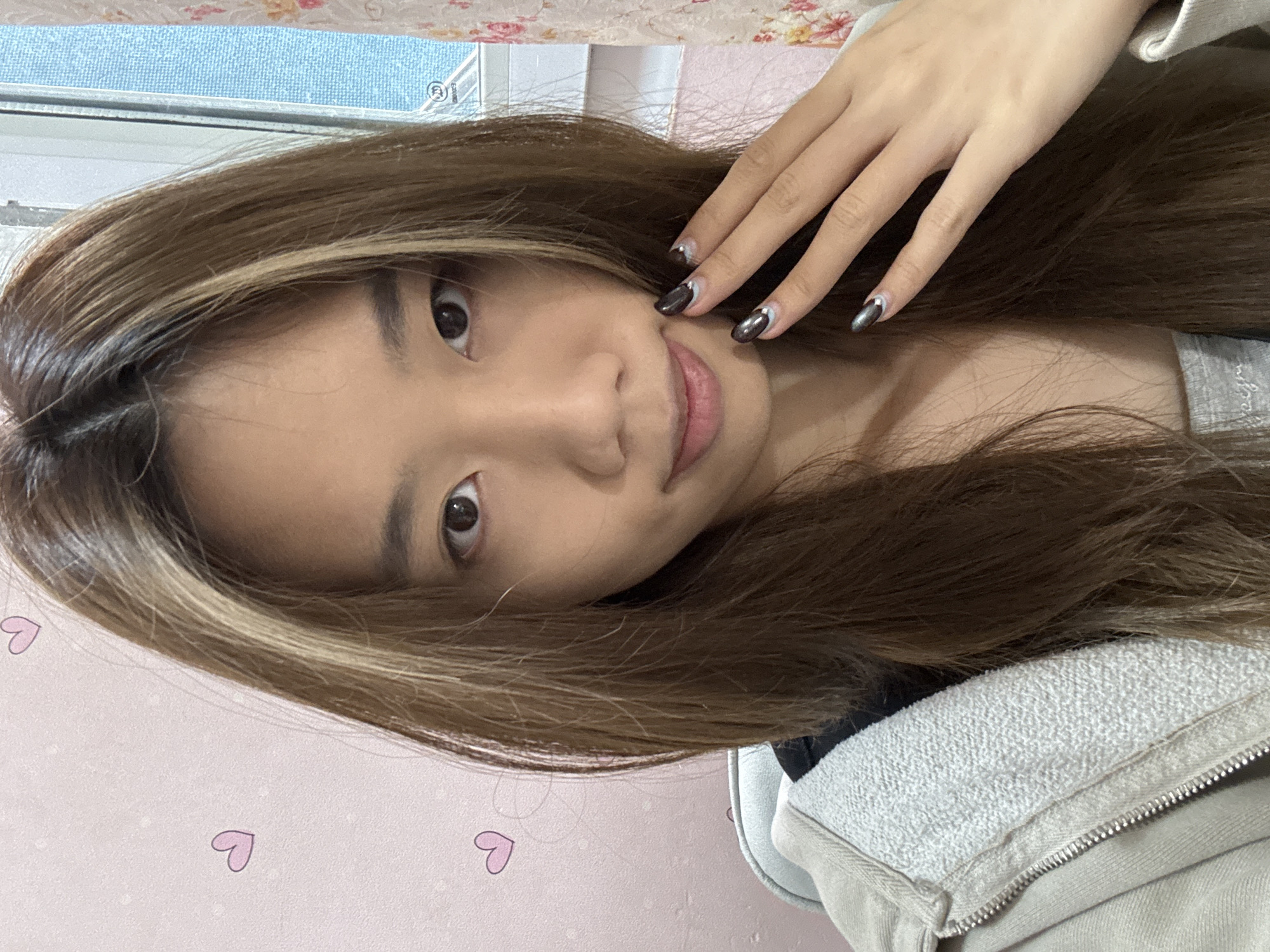 A young woman with brown hair with blonde highlights is looking directly at the camera with a slight smile. She has manicured nails with dark polish and silver accents. The background includes a pink wall with small heart patterns, a window, and part of a beige zippered jacket. The image does not provide any information regarding the location in the city of Shapingba District, China.