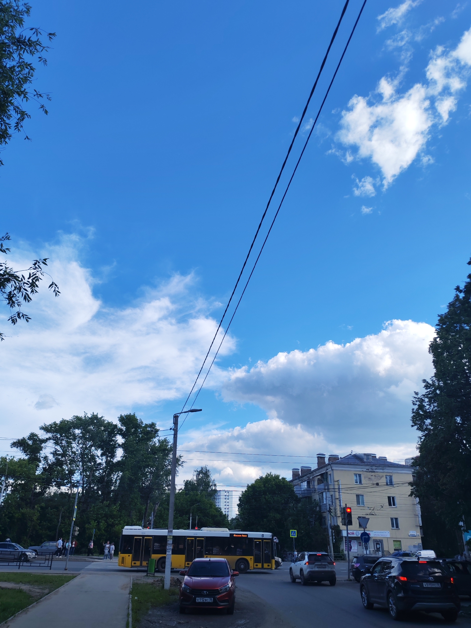 The image depicts a bustling street scene in Izhevsk, Russia, on a bright daytime, characterized by a clear blue sky dotted with fluffy white cumulus clouds. Overhead, two dark power lines stretch diagonally across the upper half of the frame.
On the left side of the street, a pedestrian crosswalk is active, with several individuals seen walking across the road. A large yellow public bus, displaying partially visible text that appears to be "Cesaul," is positioned near the crosswalk, likely stopped or in motion. A red car (license plate X 112 BC 18) is parked prominently in the foreground, with part of a dark sedan also visible near the bus. This side of the street is heavily lined with lush green trees.
On the right side, a multi-lane road is populated with several vehicles. A dark SUV (license plate A 515 PC 193) is visible from the rear, with a white SUV ahead of it. A traffic light, clearly displaying a red signal, indicates stopped traffic. Behind the vehicles and traffic light, a multi-story building with a light yellow facade, a grey roof, and multiple windows stands. This building appears to be an older urban structure, possibly housing both residential and commercial units, with some indistinct signs visible on its lower level. Trees also line this side of the street.
The scene captures a typical urban environment with active vehicular and pedestrian traffic under sunny, partly cloudy conditions.