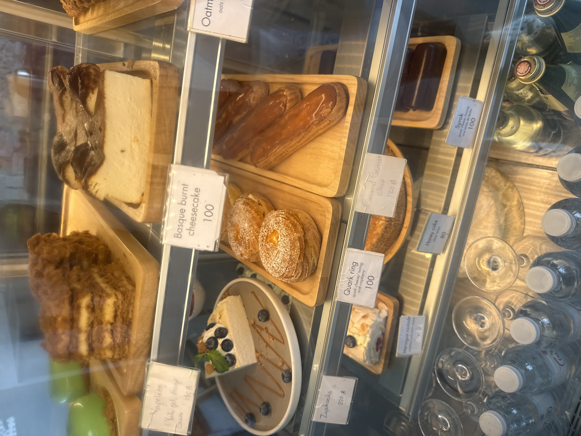 The image shows a display case filled with various desserts and beverages, likely in a cafe or bakery. In the top shelf, there is a stack of layered pastry that appears to be a Napoleon cake, a slice of green-glazed cake, and a slice of Basque burnt cheesecake. Below that, on the second shelf, are several pastries including what looks like croissants or danishes, eclairs with a caramel glaze, and a slice of cake topped with blueberries and cream. The bottom shelf displays more desserts such as a rolled cake with berries, chocolate bars, and possibly a financier cake. In front of the bottom shelf are several bottles of water and some empty glassware. A small white sign with handwritten text and prices is visible in front of many of the items. The overall setting suggests a retail environment for sweets and drinks. The presence of "Baht" (B) next to some prices indicates the currency, which further suggests a location in Thailand. The text on the signs like "Meringue roulade" and "Honey cake" suggests a menu of desserts. The mention of "Ko Samui District, Thailand" in the prompt provides geographical context, making the cafe a likely location for such a display.