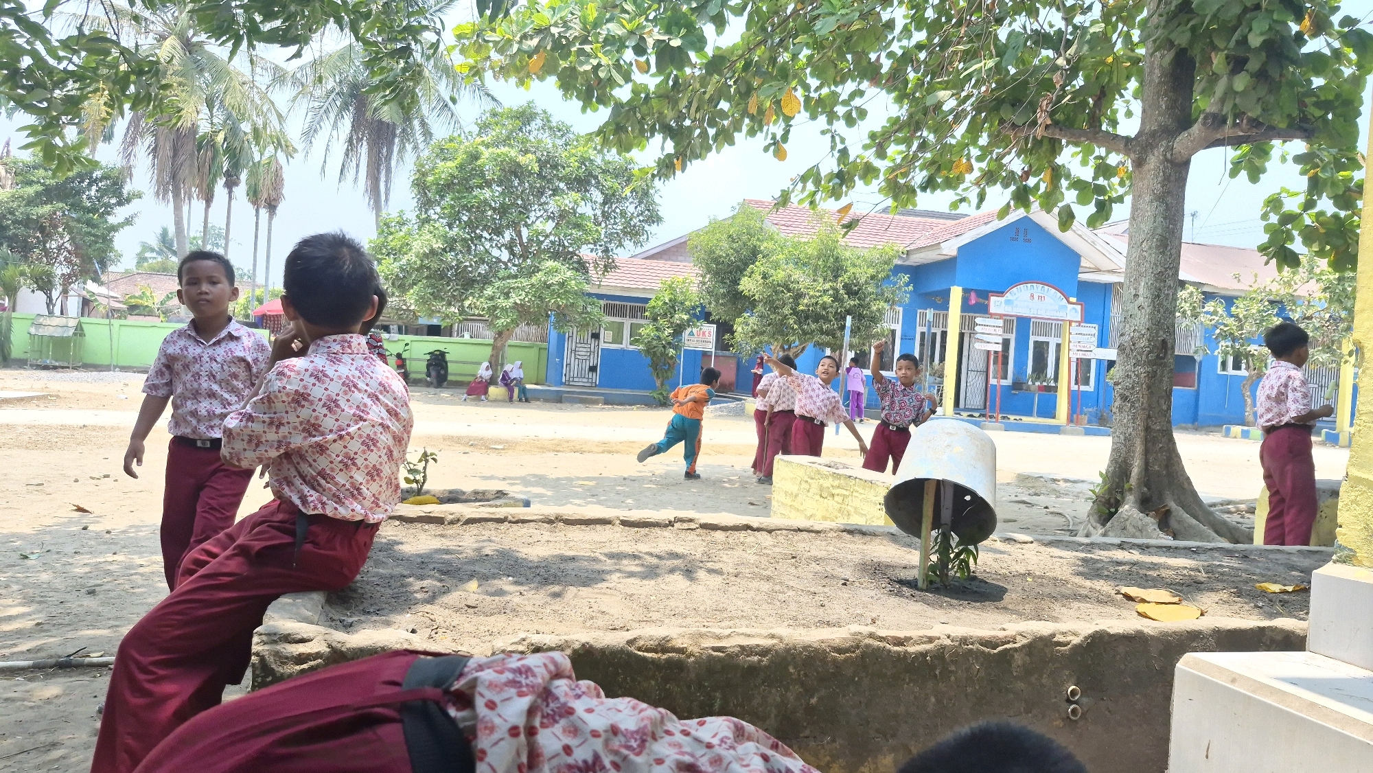 In the city of Putak, Indonesia, a group of children in school uniforms are gathered outdoors. The school building, painted bright blue with a red roof, is visible in the background. Several children in patterned shirts and maroon pants are interacting. Two boys stand near the foreground, one facing the camera and the other with his back to it. Further in the background, other children are seen playing. The environment appears to be a schoolyard with sandy ground, trees, and some vegetation. The sky is bright, suggesting daytime.