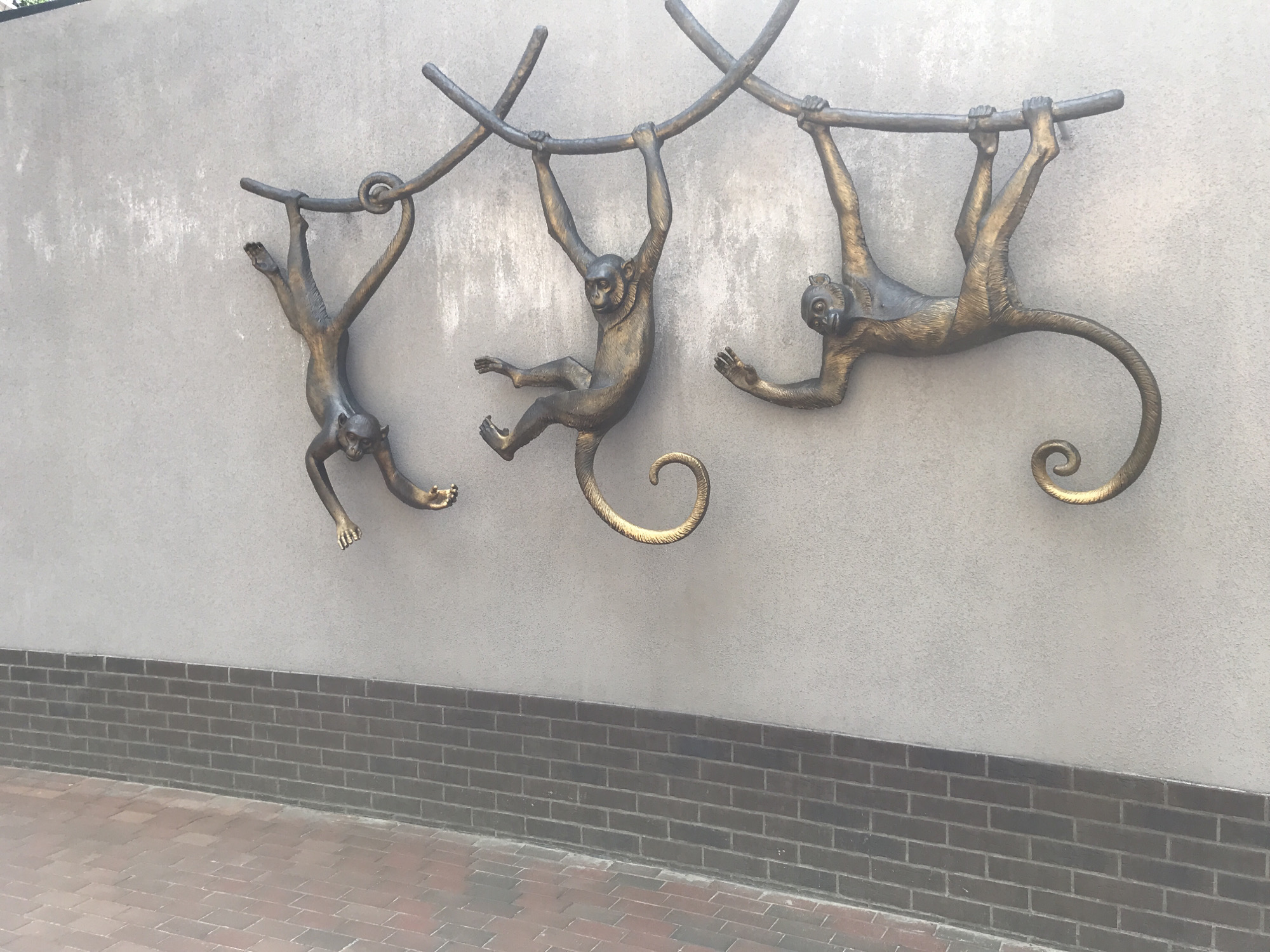 The image shows a bronze sculpture of three monkeys hanging from branches on a light gray wall. The monkeys are positioned at different angles, with one hanging upside down, one swinging, and one hanging by its hands. The branches are also made of bronze and curve across the wall. The wall has a textured finish. Below the wall is a strip of dark gray bricks, and below that is a sidewalk paved with reddish-brown bricks laid in a herringbone pattern. The lighting suggests it is daytime. The scene is likely an exterior public space in Kharkiv, Ukraine, possibly a park or a street.