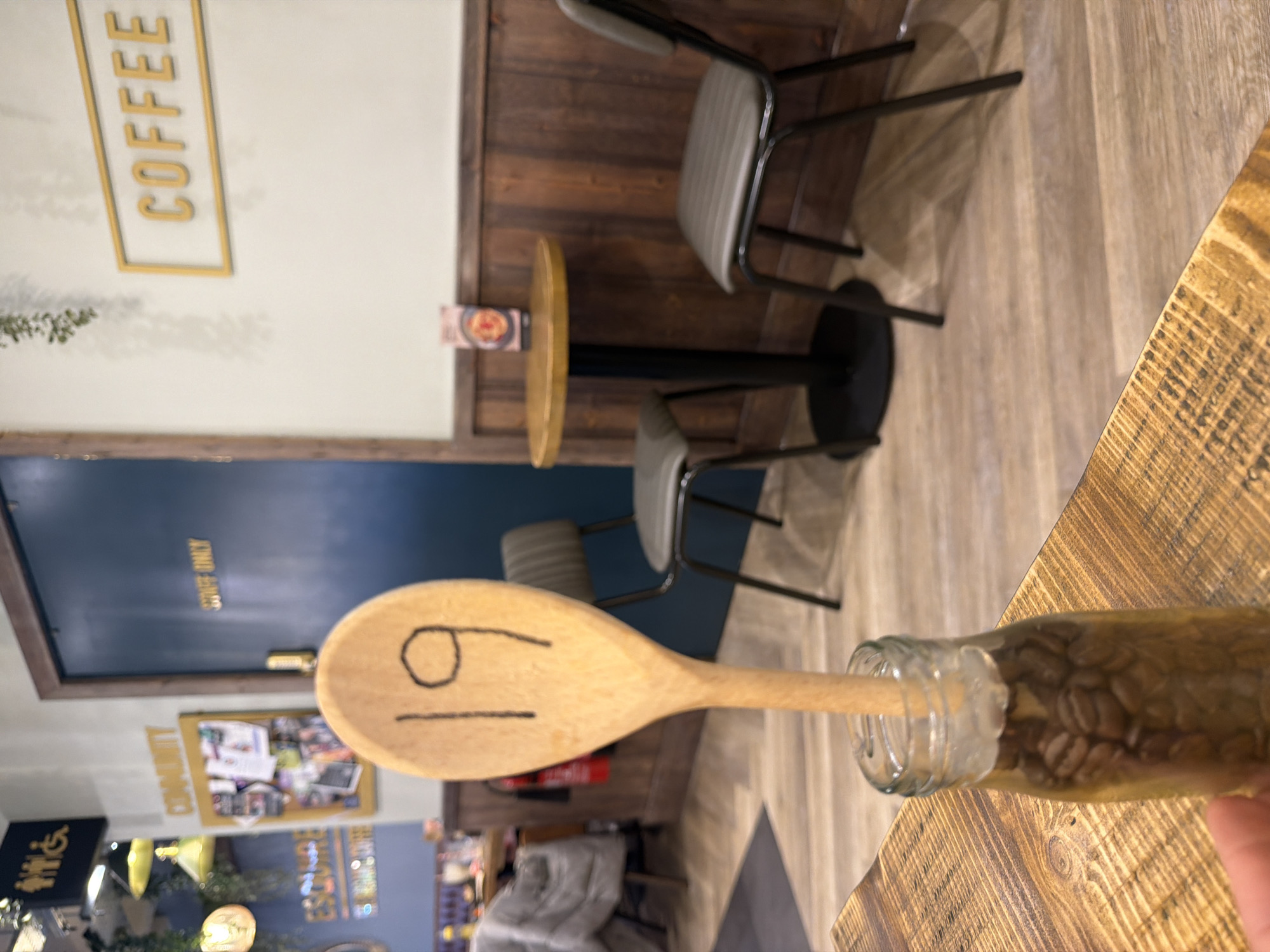 The image shows an interior space, possibly a cafe or coffee shop, with a wooden spoon inserted into a bottle filled with coffee beans resting on a wooden table. The spoon has a drawn face on it. Several chairs are positioned around small tables with a wooden countertop. In the background, there's a door with "STAFF ONLY" written on it, along with a corkboard displaying various images and announcements. A "COFFEE" sign is visible on the wall. The overall scene is rotated, so details are viewed from an unusual angle. The exact location in Mole Valley, United Kingdom, is not discernible from the image.
