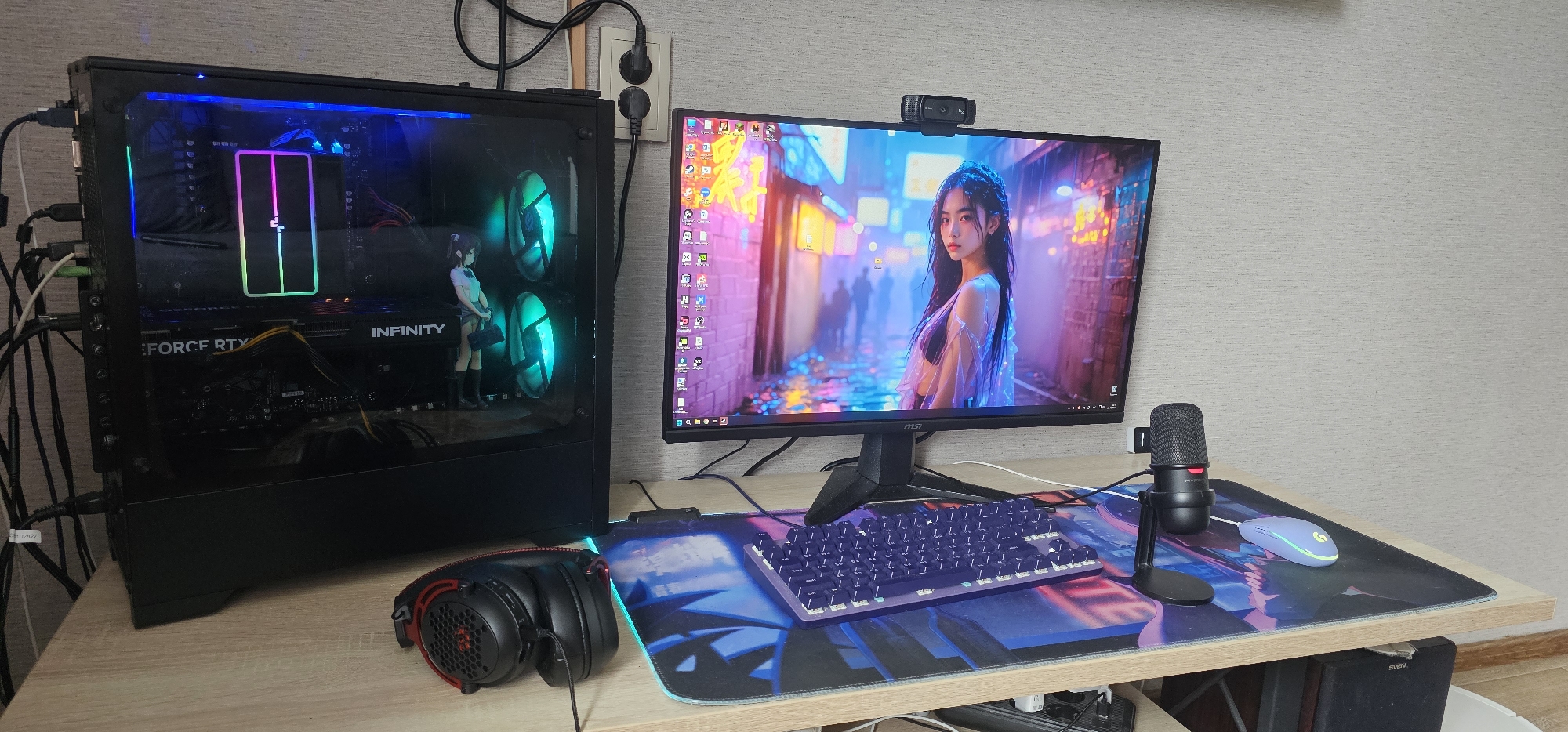 The image shows a desk setup for a computer. On the left is a black computer tower with a transparent side panel, revealing internal components and RGB lighting. A graphics card labeled "INFINITY" and "GEFORCE RTX" is visible. A figurine of a young woman in a school uniform stands inside the computer case. In front of the computer tower is a pair of black and red headphones.
In the center is a computer monitor displaying a digital artwork of a young woman in a dimly lit, neon-lit street. Above the monitor is a black webcam. Below the monitor is a purple mechanical keyboard and a large desk mat with a blue and black abstract design. To the right of the keyboard is a black microphone on a stand. To the right of the microphone is a light blue computer mouse.
The desk is a light wood laminate. In the background, a beige textured wall is visible. A white electrical outlet with two plugs is mounted on the wall to the left of the monitor. Cables are visible connected to the computer tower.
The location is described as being in Zarechny, Russia. However, there are no specific visual cues within the image to confirm this location.