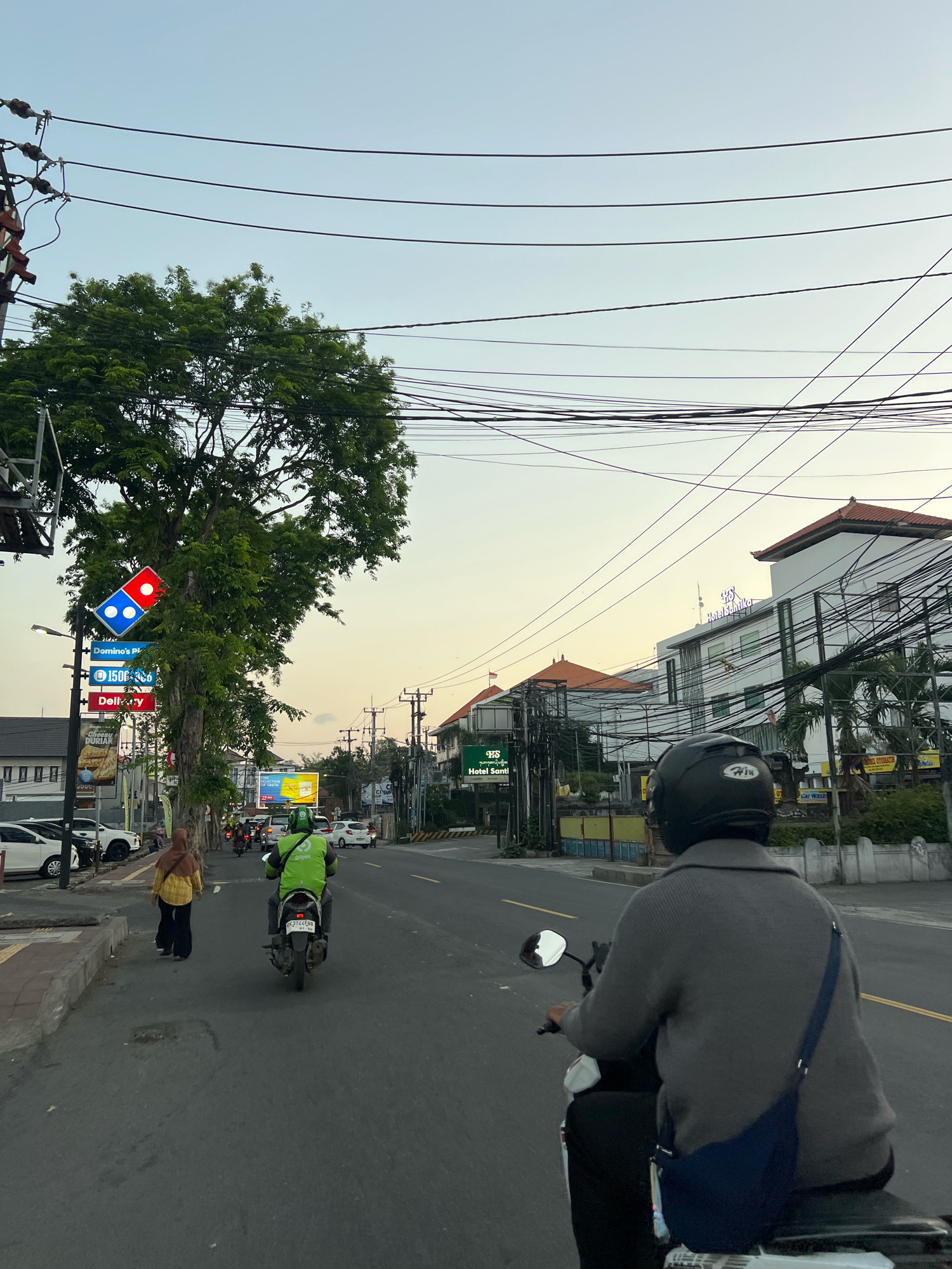 A low-angle, slightly over-the-shoulder shot captures a street scene in Kuta, Indonesia, during a warm, overcast sunset. The foreground is dominated by a person riding a scooter, their back to the camera, wearing a gray sweater and a black helmet. A dark blue sling bag is draped across their chest. The scooter's license plate is partially visible.
To the left, a woman in a yellow top and dark pants walks along a paved sidewalk. Behind her, a person on a green scooter with a Gojek logo is seen from the back, navigating the road. The street itself is asphalt with yellow dividing lines.
Several vehicles are parked along the left side of the road, including white cars. A large, leafy tree stands prominently on the left, partially obscuring a tall pole with multiple signs. One of these signs features the Domino's Pizza logo with its iconic red and blue circles. Other signs advertise "Domino's Pizza," "150 R," and "Delivery."
In the background, buildings line the right side of the street. One building, identified by signage as "Hotel Santi," is a multi-story structure with a modern white facade and dark-tiled roofs. Numerous electrical wires are strung across the sky, creating a dense network above the street and buildings. The sky transitions from a pale blue at the top to a warmer, yellowish hue near the horizon, indicative of sunset. Palm trees are visible interspersed with the buildings. The overall atmosphere suggests a bustling urban environment in the late afternoon.