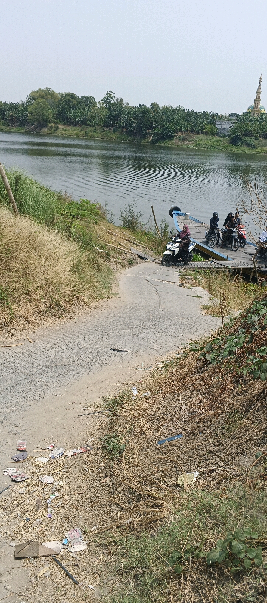 The image depicts a waterside scene, likely in Brumbun, Indonesia. A dirt and asphalt path slopes down towards a wooden dock extending into a body of water. Several motorcycles are parked on the dock, and people are visible riding them. Some of the motorcycles are positioned on a ramp leading onto the dock. The water has ripples, suggesting a slight breeze or movement. The surrounding environment consists of dry, tall grass on the banks and lush green vegetation, including what appear to be banana trees, further in the distance. On the far side of the water, a building with a dome and a tall, slender tower, resembling a mosque, is visible. The ground near the path is littered with various pieces of trash, including wrappers and paper. The sky is overcast and pale.