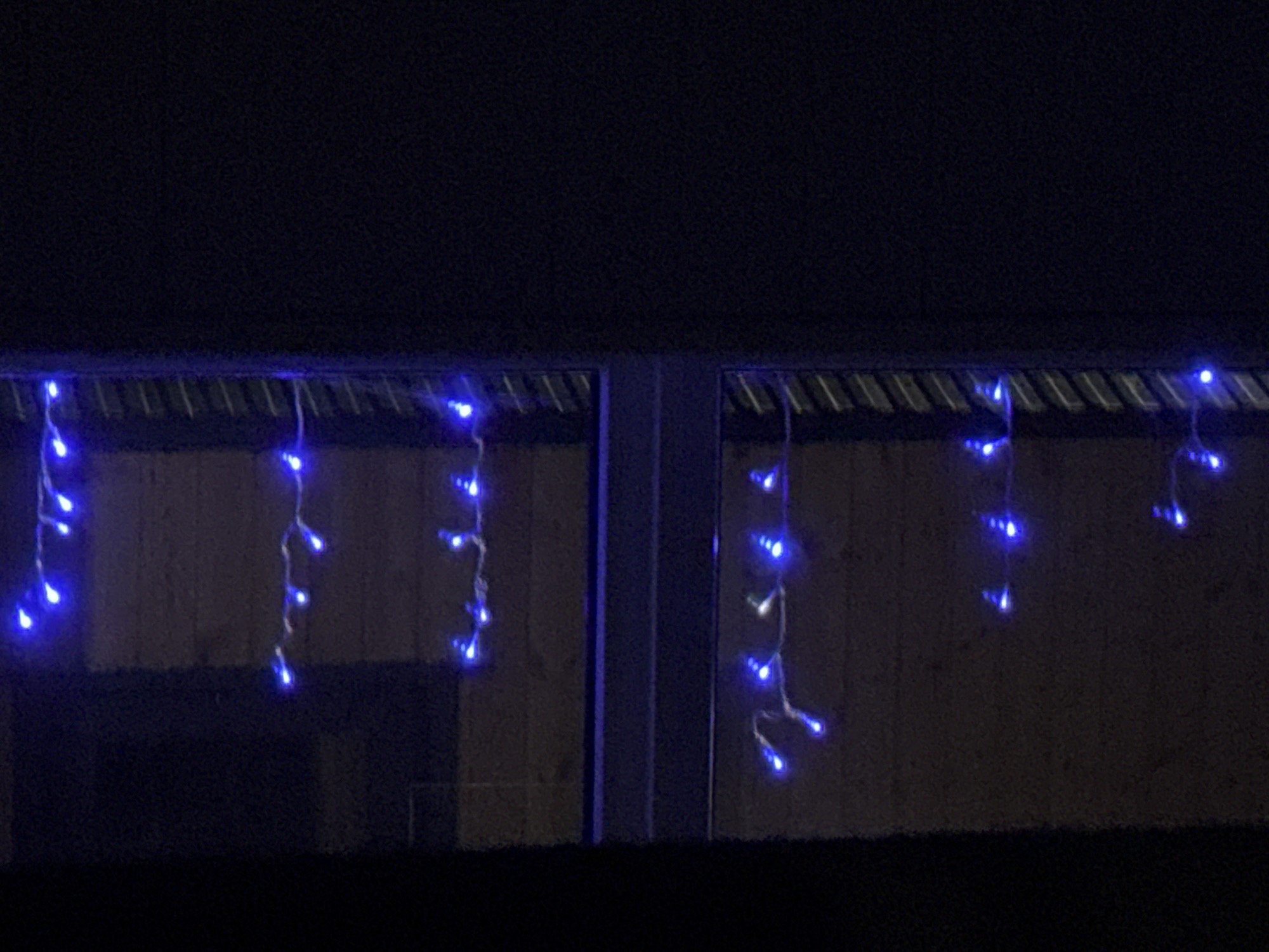 The image shows a nighttime view of a building with blue icicle-style string lights draped across the top of two windows, with some lights hanging down. The building appears to be a single-story structure, possibly a house or a small commercial establishment. The lights are spaced out and illuminate the facade of the building, creating a festive atmosphere. The background is dark and indistinct, suggesting the photo was taken in an outdoor setting at night. There are no people or visible signs of activity in the image. The overall impression is one of a quiet, decorated exterior.