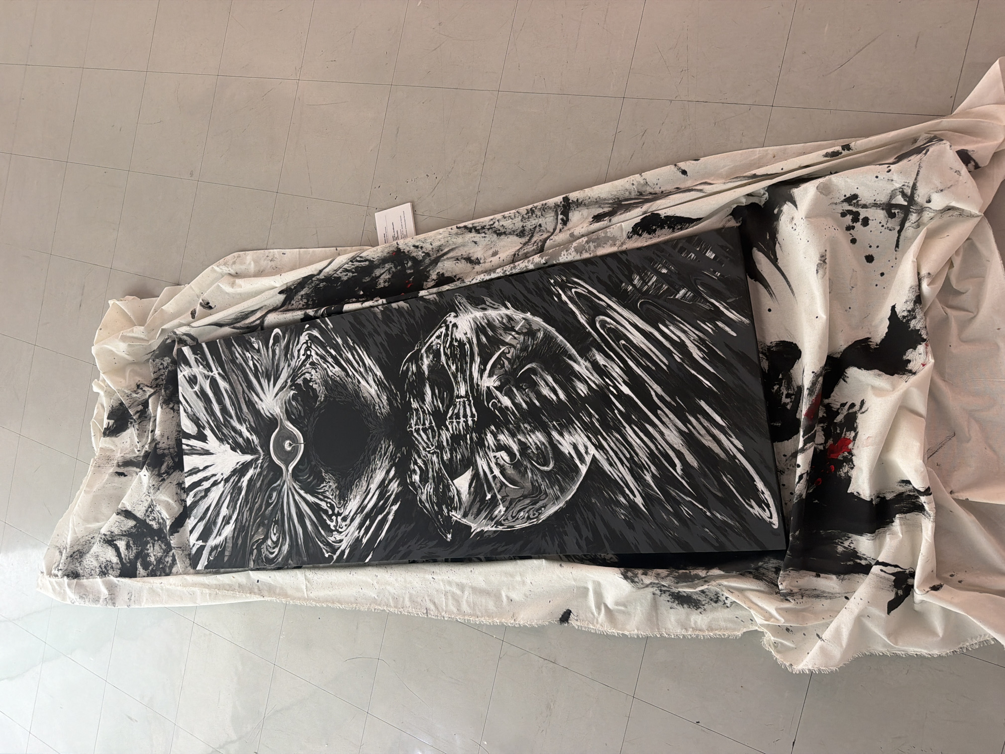 An abstract painting is laid out on a white cloth with black and red paint splatters on the floor. The painting is black and white with dynamic brushstrokes creating an illusion of depth and movement. The white cloth is draped around the painting, also showing signs of paint splatters. The floor is tiled in a light grey color. There is a small white tag with text on the cloth near the top right of the painting. No people or specific city landmarks are visible in the image. The scene is indoors with artificial lighting.