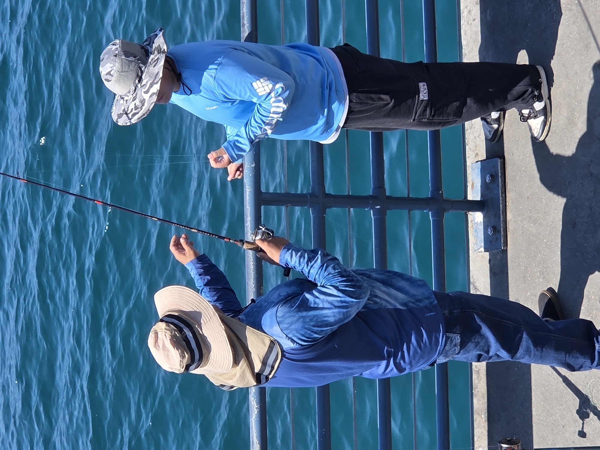 Two individuals are fishing from a pier in Santa Monica, California, on a sunny day. The ocean is a vibrant turquoise blue, with visible ripples. The pier has a dark blue railing with metal supports.
The individual on the left is wearing a wide-brimmed, light-colored hat with a dark band and a neck shade. They have on a long-sleeved, light blue shirt with a patterned design and dark blue jeans. They are holding a fishing rod and appear to be adjusting the line or reel.
The individual on the right is wearing a camouflage-patterned hat and a long-sleeved, bright blue shirt with white lettering on the sleeve. They are wearing black cargo pants. This individual is also holding a fishing rod, with their hands near the reel.
The ground of the pier is a light gray concrete surface, with shadows from the individuals and the railing visible.