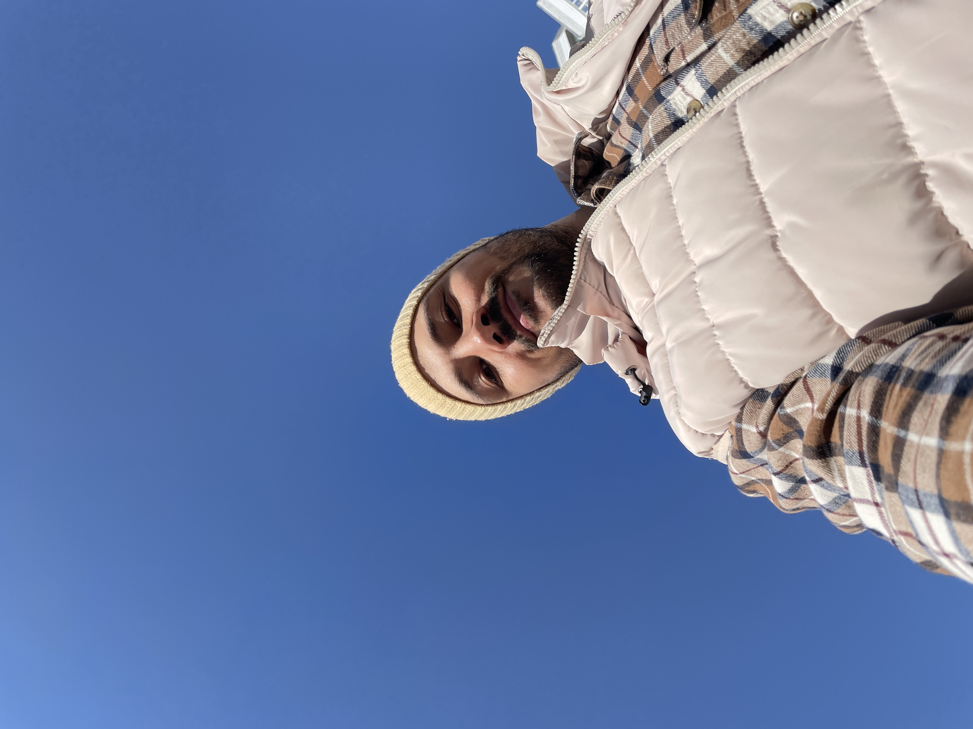 A man wearing a beige beanie, plaid shirt, and puffy vest is seen from a low angle against a clear blue sky. The location cannot be determined from the image alone.