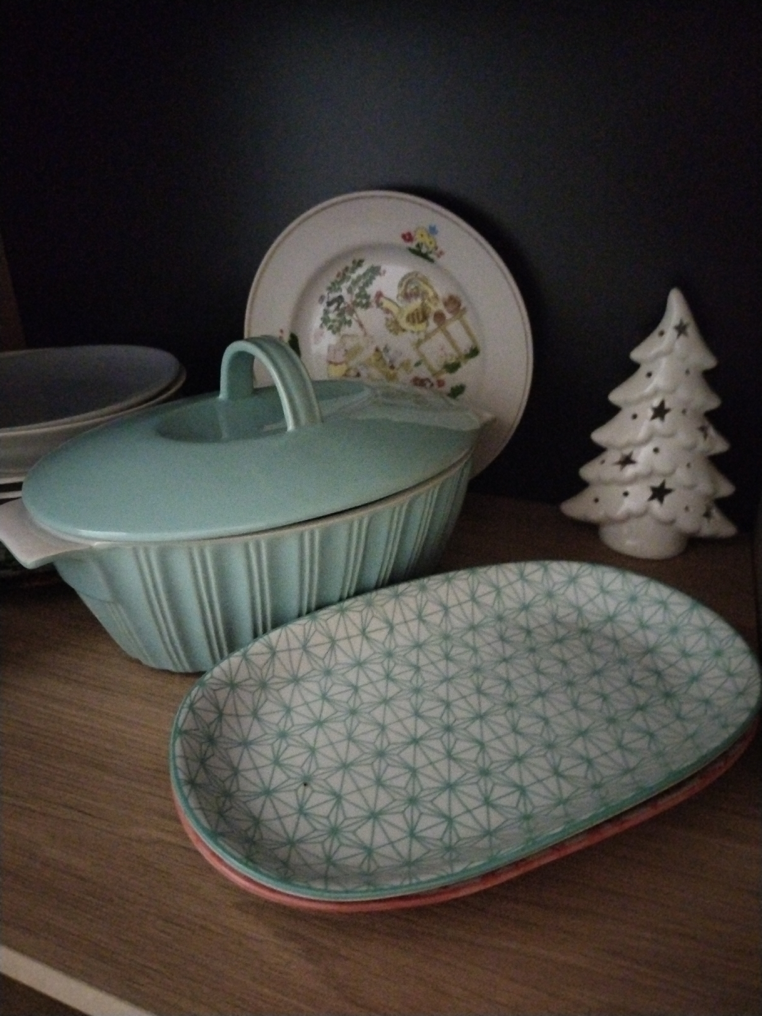 An interior scene located in Бараўлянскі сельскі Савет, Belarus, displays various ceramic dishes and a decorative figurine arranged on a light brown wooden surface against a dark, featureless background.
In the mid-background, a circular, cream-colored ceramic plate stands upright, featuring a multi-colored illustration of figures and foliage. To its right, a white, ceramic, three-tiered Christmas tree figurine with multiple star-shaped perforations is positioned. Partially visible on the far left, in the background, is the rim of a grey circular dish.
In the foreground, an oval-shaped, lidded, light blue-green ceramic serving dish, characterized by vertical fluting on its exterior and an integrated loop handle on its lid, rests on the wooden surface. Beneath its front edge, a small white border, possibly of another dish, is visible. Adjacent to the serving dish, a stack of two oval-shaped ceramic plates is present. The top plate is white with a repeating geometric pattern of interconnected light blue-green triangles. The bottom plate reveals a reddish-orange underside. The wooden surface displays a visible grain texture.