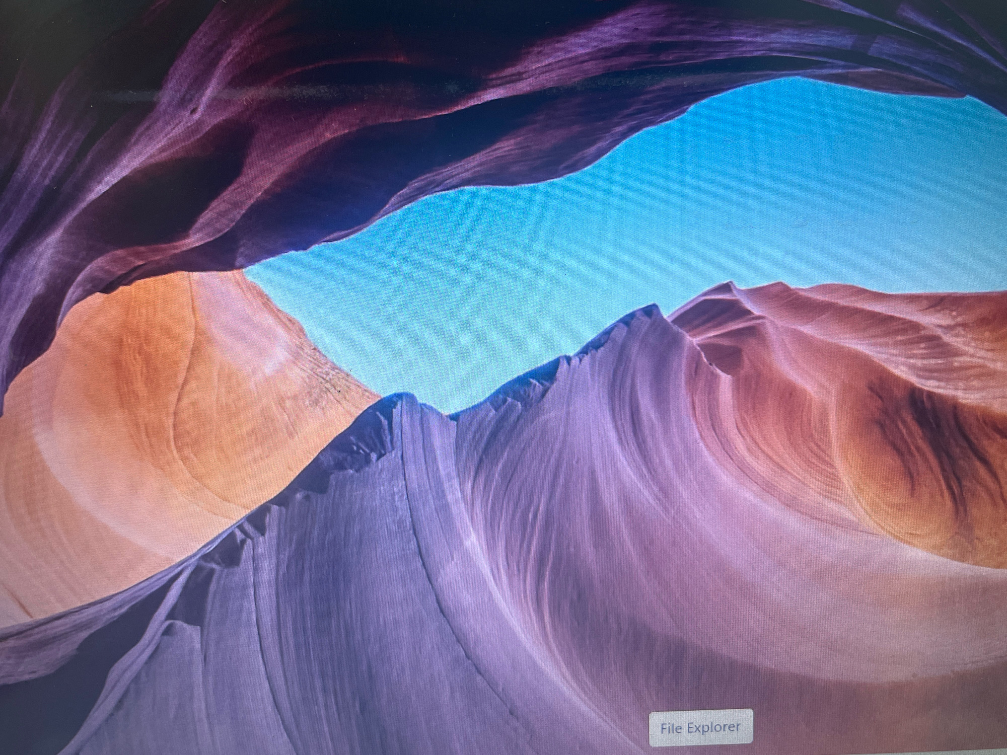 The image displays a desktop background featuring a landscape with sculpted rock formations. The dominant colors are shades of purple, orange, and blue. The sky is a bright, clear blue, while the rock formations exhibit smooth, flowing lines in hues of orange and purple. In the lower right corner of the image, a label reads "File Explorer". There are no people or discernible urban elements present. The scene depicts a natural geological formation, possibly Antelope Canyon in Arizona, USA, rendered as a digital image on a screen. The prompt's request to include the location in Manesar, India, cannot be fulfilled as the image does not provide any information to indicate it is located there, nor does it depict any recognizable features of that city.