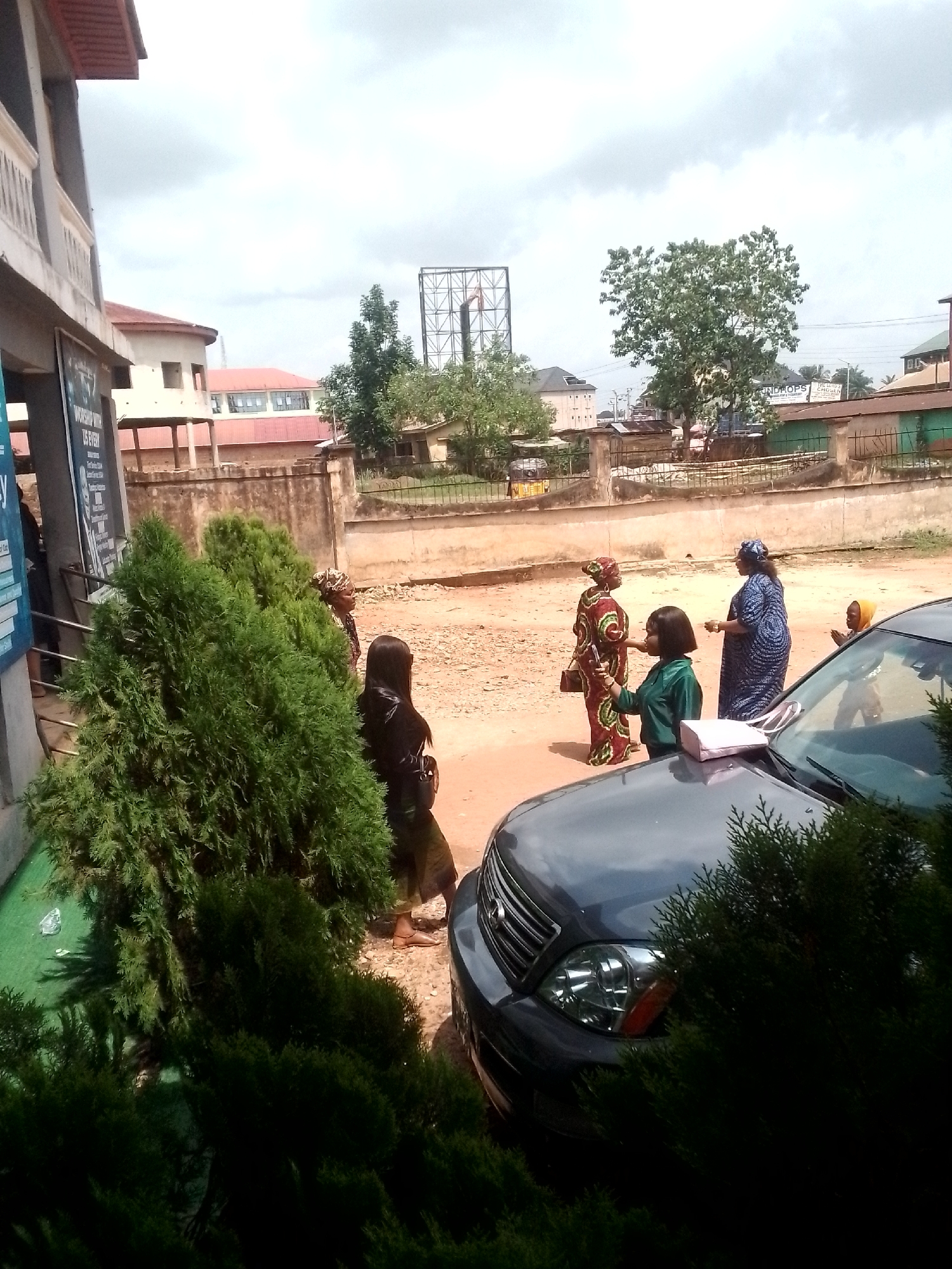The image depicts a street scene in Orlu, Nigeria. Several women are congregated in the street near a parked dark grey SUV. One woman is wearing a patterned dress and carrying a purse; another is dressed in a blue patterned robe. A child with a yellow head covering stands near the car, partially obscured. Another woman in a green top is interacting with the woman in the patterned dress. A building with a sign is visible on the left, partially obscured by trees. Across the street is a low wall and various buildings and trees. A billboard is visible in the distance. The ground appears to be reddish-brown. The sky is overcast.