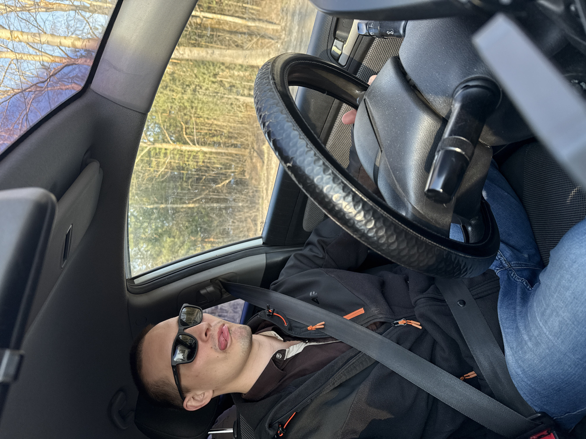 A medium-angle, interior shot depicts a man seated in the driver's seat of a car. The man is wearing a black jacket over a brown collared shirt, blue jeans, and black sunglasses. He has a seatbelt fastened across his chest. His head is tilted back against the headrest, and his mouth is slightly open, with his tongue visible. The steering wheel is positioned in front of him, and the car's dashboard and windshield are visible, revealing a wooded area outside. The location and city information provided are inconsistent with the visual content of the image, so no further location-specific information can be extrapolated.