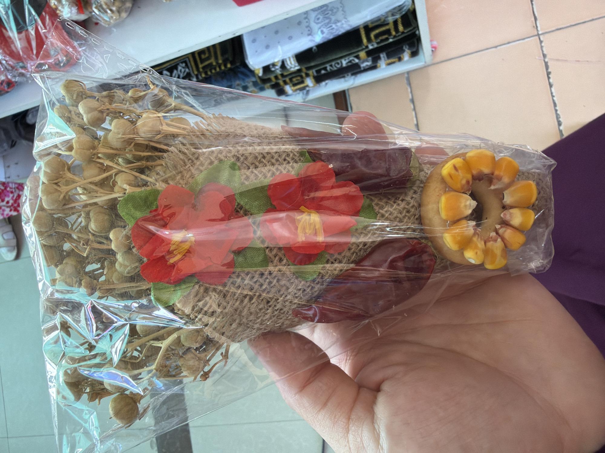 A hand holds a dried floral arrangement wrapped in clear plastic. The bouquet is made of dried seed pods on thin stems, interspersed with two red artificial flowers and a green artificial leaf. The base of the bouquet is wrapped in burlap. At the bottom of the bouquet, a ring of corn kernels is attached to a light brown circular object, also wrapped in plastic. The background shows a tiled floor and shelves displaying fabric rolls and other packaged goods, indicative of a shop or market stall in Zangiata, Uzbekistan.