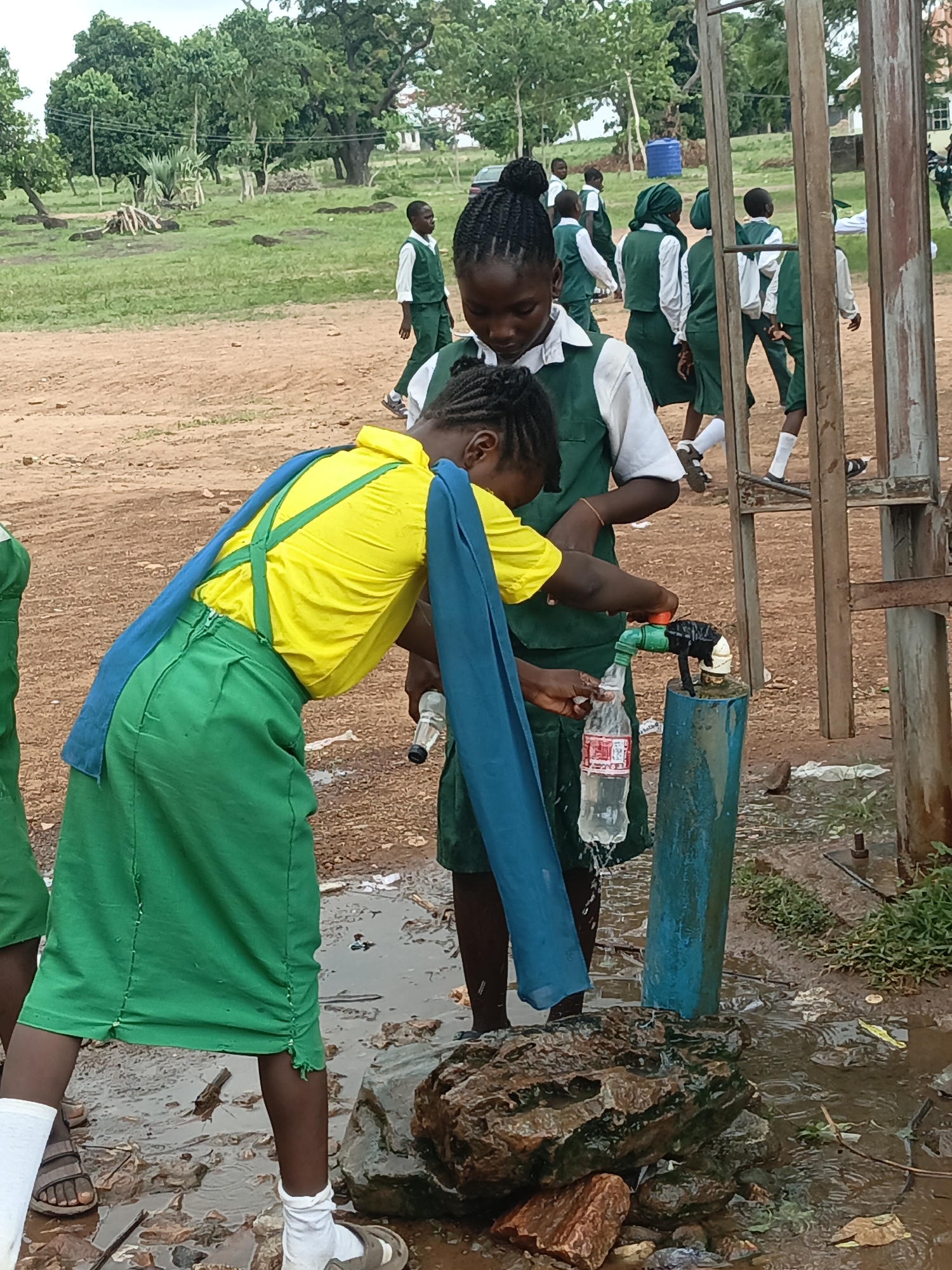 Two schoolgirls in Akwanga, Nigeria are filling water bottles at an outdoor tap. One girl wears a yellow shirt, green skirt, and blue sash, while the other wears a white shirt and green skirt. The tap is attached to a blue pipe. Other students in green and white uniforms are visible in the background. The ground around the tap is muddy and littered. A gate structure is visible in the background. Trees and grass are also visible in the distance.
