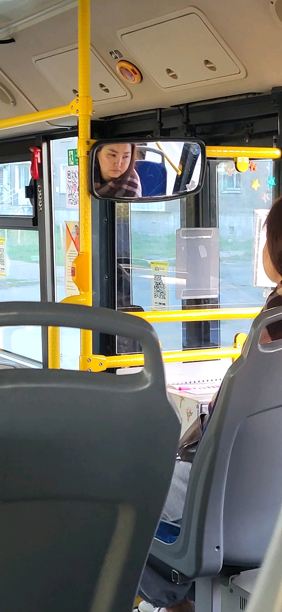 The image is a medium shot taken inside a bus, focusing on the interior from the perspective of a seated passenger. In the foreground, the backs of two gray bus seats are visible. A yellow pole runs vertically to the left of the frame, with yellow horizontal bars extending across the bus. A side-view mirror is mounted on the right side of the yellow pole. Reflected in the mirror is the face of a woman with dark hair, wearing a plaid shirt. She appears to be looking downwards. Outside the bus windows, buildings and greenery are faintly visible. The environment appears to be a public transportation vehicle, likely in Oskemen, Kazakhstan, given the context of the prompt. QR codes are visible on some of the window advertisements. The lighting suggests it is daytime.