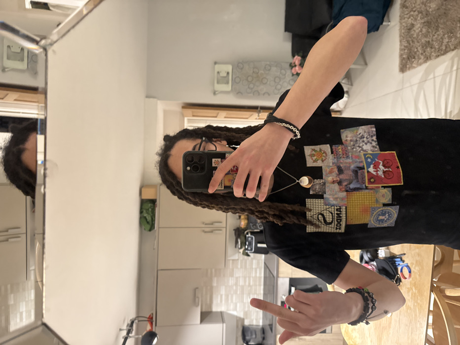 A person with long dreadlocks and glasses takes a mirror selfie in a kitchen. They wear a black t-shirt with several graphic patches on it. The phone has a black case and stickers on it. The person wears bracelets on both wrists and a necklace. Their middle finger is raised in the air. The kitchen has white cabinets and subway tile backsplash. A reflection of the person is visible in the mirror to the left.