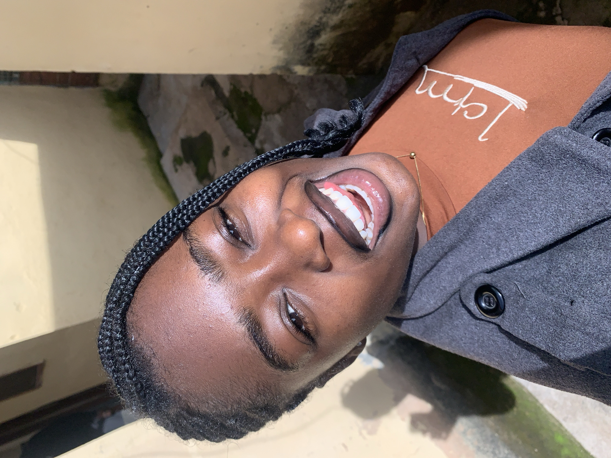 An outdoor, eye-level, close-up shot shows a young Black woman with braided hair smiling with her tongue sticking out. She is wearing a dark gray coat over a rust-colored t-shirt with white cursive writing on it. The background shows a building and a pathway. The lighting suggests it is daytime. This location is in New Stadium, Nigeria.