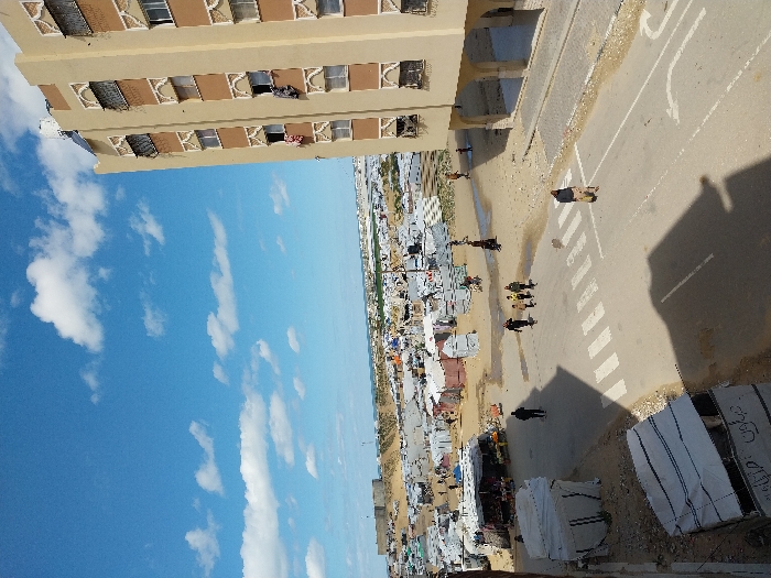 An elevated view captures a sprawling, densely populated scene in Khan Yunis, Palestinian Territory, under a blue sky with scattered white clouds. To the right, a multi-story, light beige building with brown decorative panels and arched motifs features numerous windows, some open with laundry hanging out. Solar panels and water tanks are visible on its roof. In the mid-ground, a paved road with a white-striped crosswalk and a right-turn arrow shows the distinct shadow of a nearby structure. One individual, wearing a dark jacket and light trousers, walks alone on the right side of the road. A group of four people, two adults and two children, walks away from the viewer on the left side of the road, having crossed the pedestrian marking. To the left of the road, two temporary structures made of white tarpaulin draped over frames, possibly market stalls or shelters, stand on unpaved ground. The vast background is dominated by an extensive, makeshift encampment covering a sandy, dusty landscape, consisting of countless temporary shelters, including white, blue, and grey tents, stretching towards the horizon where more permanent structures are faintly visible. Sparse human activity is observed within the encampment.