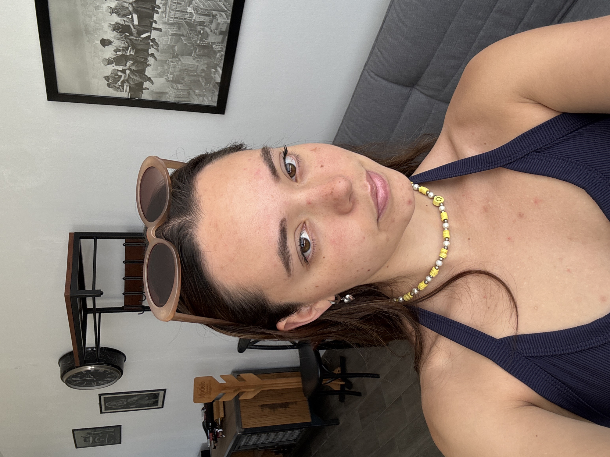 A woman is facing the camera indoors. She wears sunglasses perched on her head and a navy blue tank top. She is wearing a yellow and silver beaded necklace with smiley face beads. A black and white framed photograph hangs on the wall behind her. In the background are a grey couch, a metal and wood rack, and a clock.
