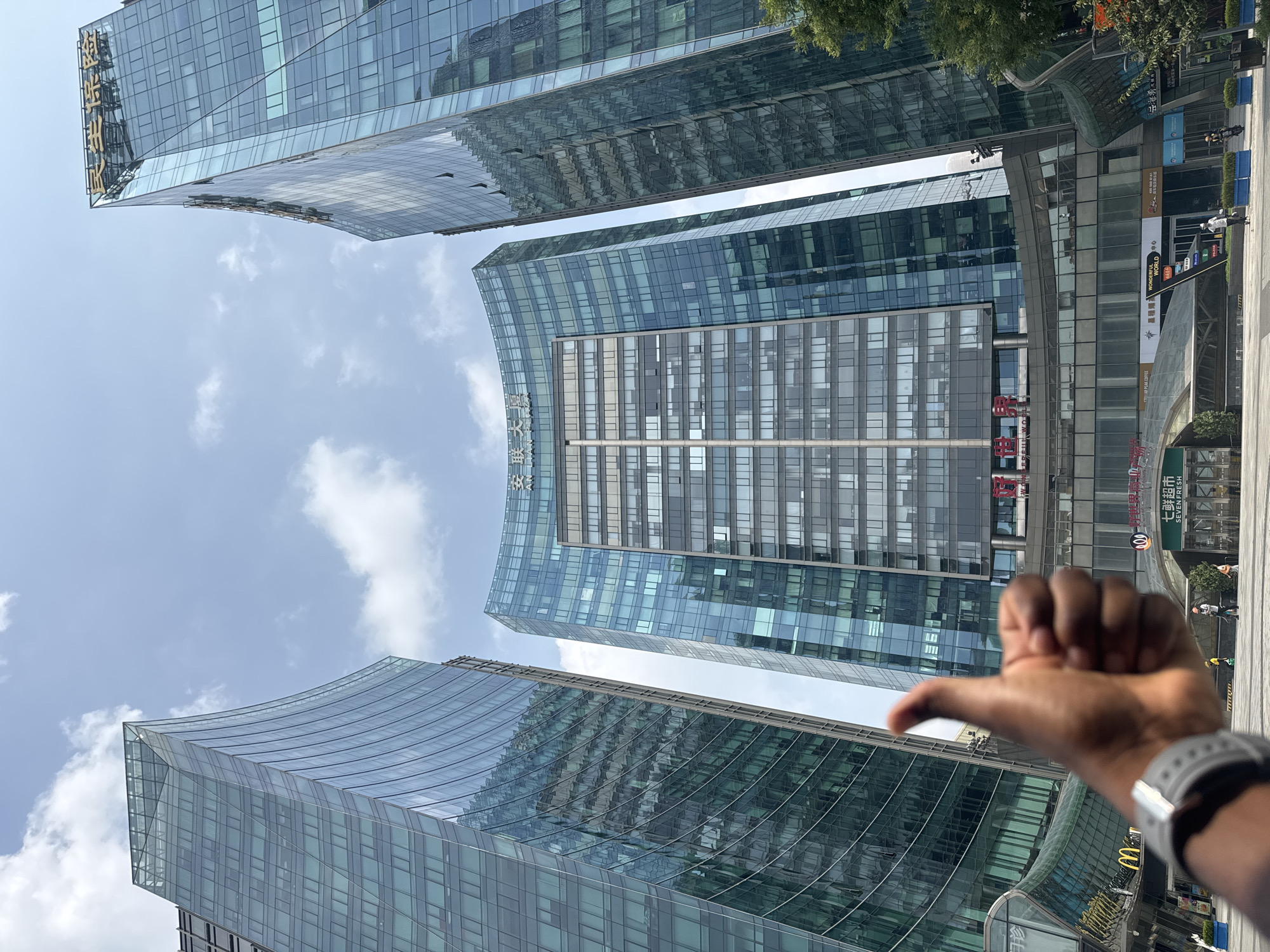 In a low-angle outdoor shot, three modern glass skyscrapers dominate the frame, with a clear blue sky dotted with white clouds overhead. The buildings feature curved and angular designs, their facades reflecting the sky and surrounding structures. In the foreground, a person's hand is visible, giving a thumbs-up gesture. Below the skyscrapers, the ground level of the buildings appears to house various shops and businesses, including signs for "McDonald's" and "SEVEN FRESH." Several individuals are walking or standing on the ground level. The scene is likely set in the Chaoyang District of China, a business and commercial hub known for its contemporary architecture.