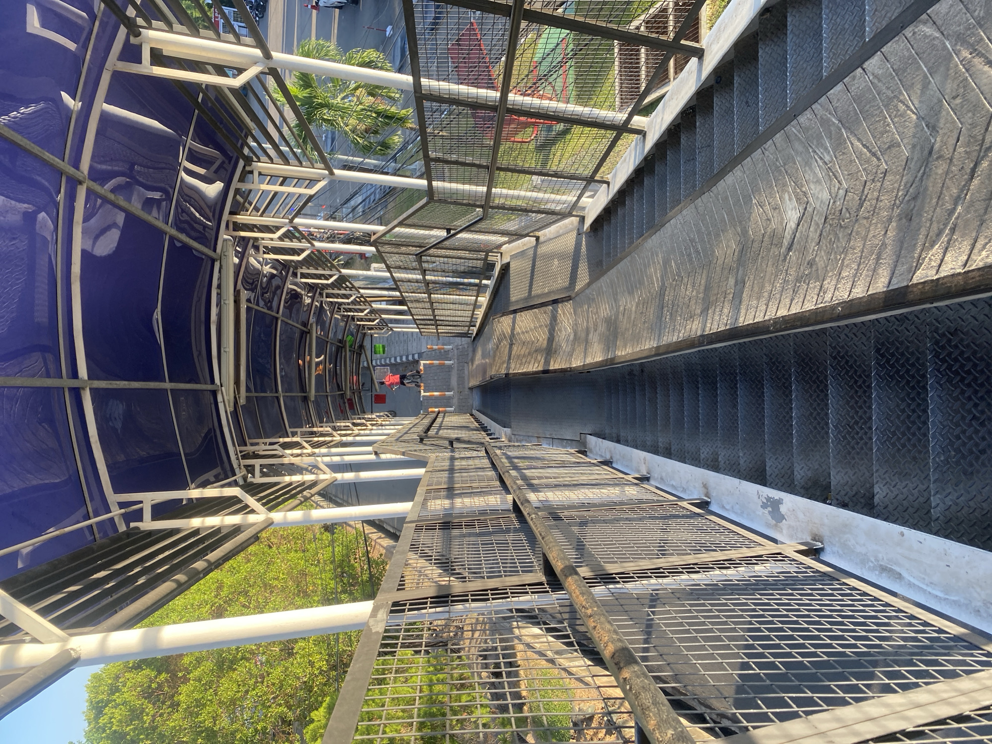 The image depicts a pedestrian overpass in Banjarbaru, Indonesia, with a view looking down a long ramp and staircase. The overpass is covered by a blue, translucent canopy supported by white pillars and metal framing. On the left side of the overpass, a wire mesh fence runs along the edge, with lush green trees visible beyond it. On the right side, a similar fence borders the structure, and beyond it, a palm tree and some buildings can be seen, along with a glimpse of a street with parked motorcycles. A person in a red shirt is visible further down the ramp, appearing to be walking or cycling. The staircase is constructed from metal treads, and the ramp features a textured surface arranged in a chevron pattern. The scene is bathed in sunlight, casting shadows across the overpass.