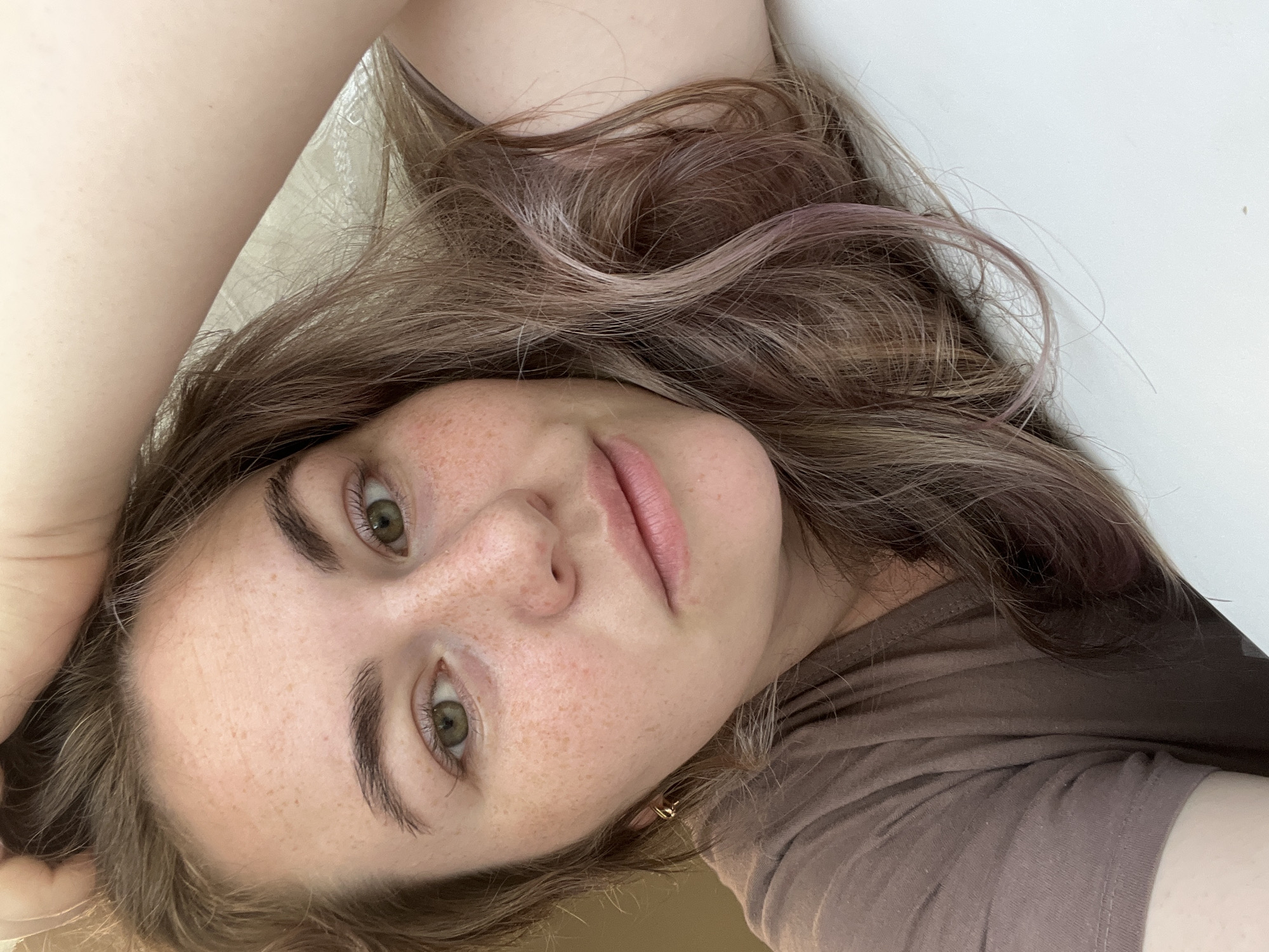 A young woman with light brown hair, freckles, and green eyes is shown in a close-up, selfie-style photograph. She is wearing a brown t-shirt. Her left hand is resting on her head, and her hair is slightly wavy with some strands of pinkish-purple color visible. The background is blurred, suggesting an indoor setting, possibly a bedroom or living space, with natural light filtering in from the right side. The woman appears relaxed. There is no specific indication of the city of Chaikovsky, Russia, in the image.