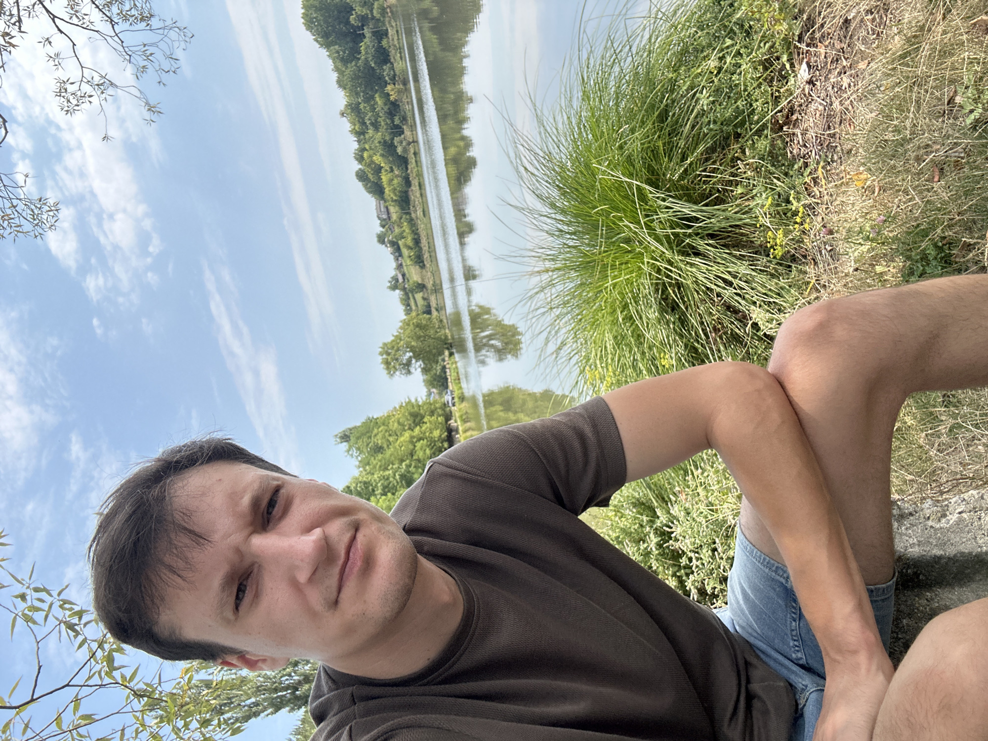 A man is taking a selfie outdoors, seated next to a body of water. He is wearing a dark, textured t-shirt and denim shorts. His right arm is bent and resting on his knee, with his left arm and hand visible as well. In the background, a tranquil lake reflects the trees and the sky. The terrain around the lake is covered in green grass and vegetation, with a distant line of trees and some buildings on a hillside. The sky is blue with scattered white clouds, and branches of a tree frame the top of the image. The scene appears to be a serene, natural setting, possibly on the outskirts of Makiv, Ukraine, given the information provided about the location.