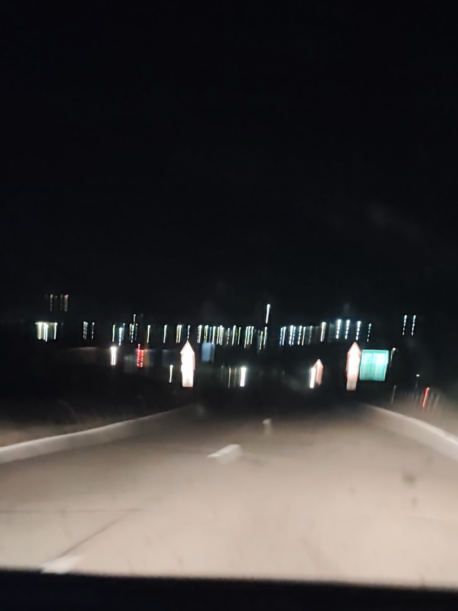 A low-resolution image shows a night scene from inside a vehicle, moving along a road. In the distance, multiple bright vertical lights or reflections are visible along what appears to be a body of water. Road markings are faintly visible on the road ahead. A green sign appears blurred on the right side of the frame. The surrounding environment is dark. No people are visible in the image. The location is specified as Belo Pole, Bulgaria.