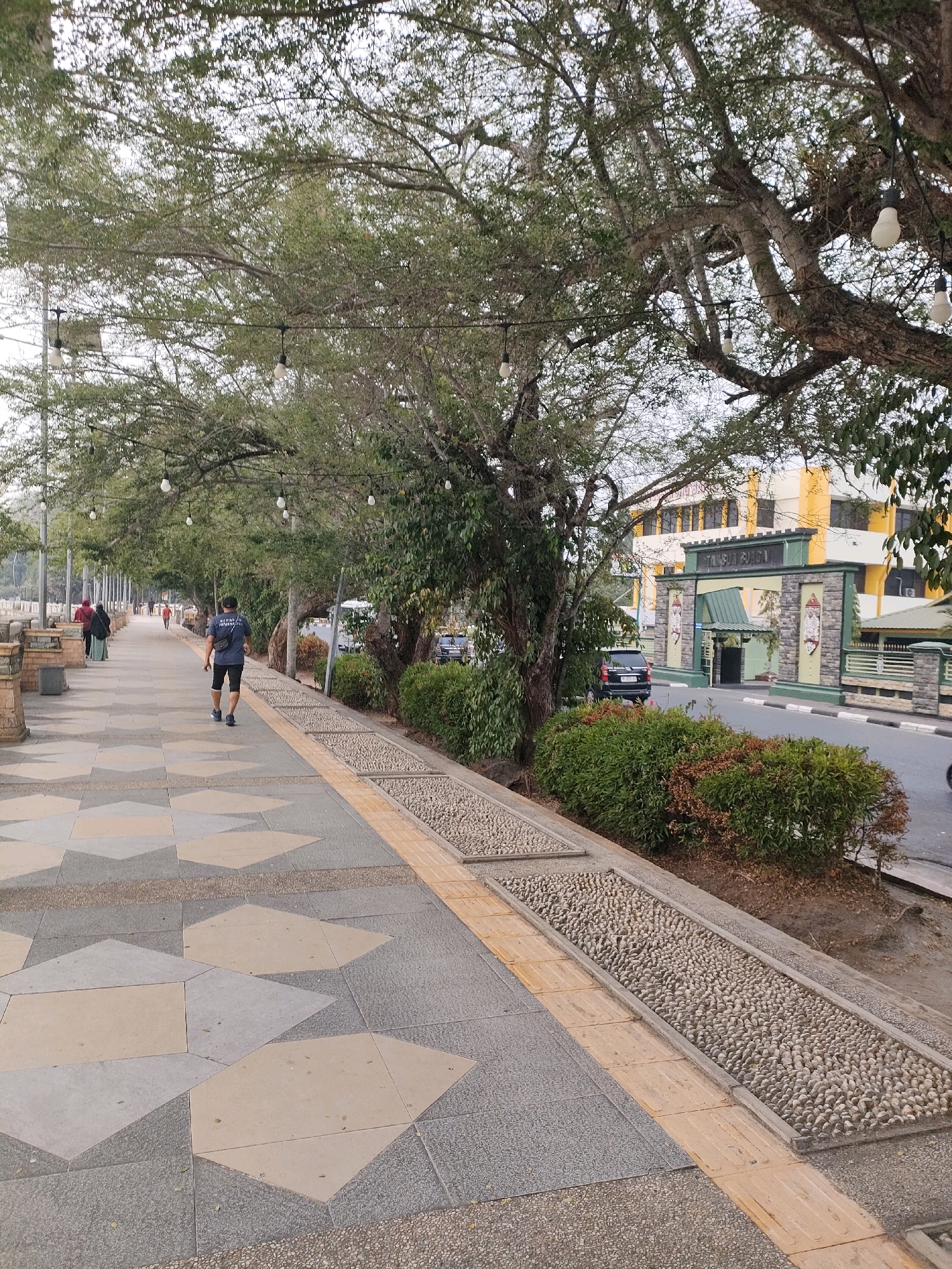 This image depicts a pedestrian walkway in Banjarmasin, Indonesia, likely along a waterfront or a main road. The walkway is paved with a decorative pattern of geometric tiles in shades of grey, beige, and a speckled dark grey. Adjacent to the tiled path is a narrow strip of gravelly texture, followed by a section filled with small, smooth pebbles, possibly for reflexology. This pebble-lined area is bordered by a yellow tactile paving strip.
To the right of the walkway, there is a green hedge and then a road with a black SUV parked near an entrance. The entrance features a stone archway with decorative panels and the inscription "TALAN SUGUL GUNA". Behind the entrance, a yellow and white building is visible.
The walkway is shaded by a canopy of large, mature trees with sprawling branches. String lights are suspended between the trees, suggesting the area may be lit at night. Several people are walking along the path, including a man in the foreground wearing a dark t-shirt and black shorts, with a dark bag slung over his shoulder. Further down the path, other individuals are seen walking in the same direction, some wearing headscarves. The background shows more trees and a hazy, overcast sky. The overall scene suggests a public area designed for recreation and passage within the city.