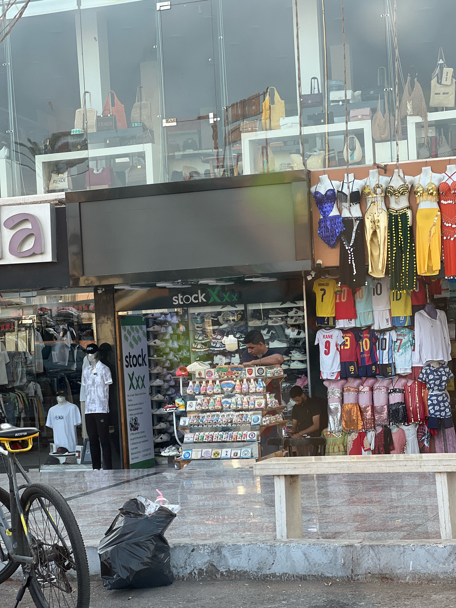 The image depicts a street-level view of storefronts in Sharm El Sheikh, Egypt. The primary focus is on a shop with a large glass facade displaying various merchandise. Inside, shelves are stocked with what appear to be shoes and clothing. Mannequins are used to display shirts and athletic wear, including soccer jerseys with names like "RONALDO" and "KANE" visible. Some mannequins are adorned with sparkly, colorful outfits, possibly costumes. A man is visible behind a counter, appearing to be attending to business. Outside the shop, a black garbage bag rests on the ground next to a bicycle. The storefront signage indicates "stock XXX". Reflections in the glass reveal more items, including handbags, and another section of the store appears to sell women's clothing. The environment suggests a commercial area.