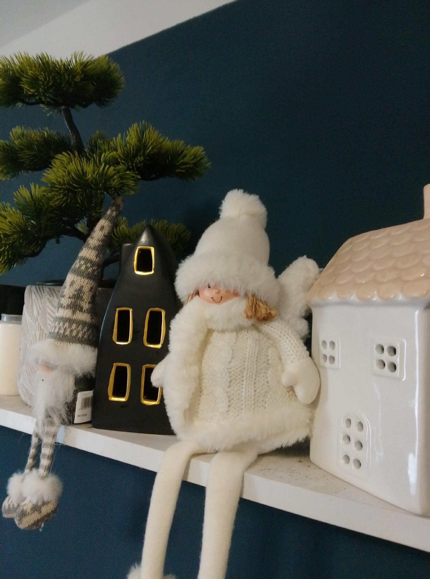 The image shows a shelf with decorative Christmas-themed items. On the left, there is a small gnome figure with a long hat and beard. Next to it is a black, house-shaped ceramic decoration with illuminated windows. To the right of that, an angel doll with a white fuzzy hat and knitted sweater is seated, with its legs dangling. Further to the right, a white ceramic house decoration with a textured roof and windows is visible. Behind the decorations, a part of a small artificial tree with green foliage can be seen. The background is a dark teal wall. The image appears to have been taken indoors, possibly in a home in Wedel, Germany, although specific details of the location cannot be determined from the image alone.