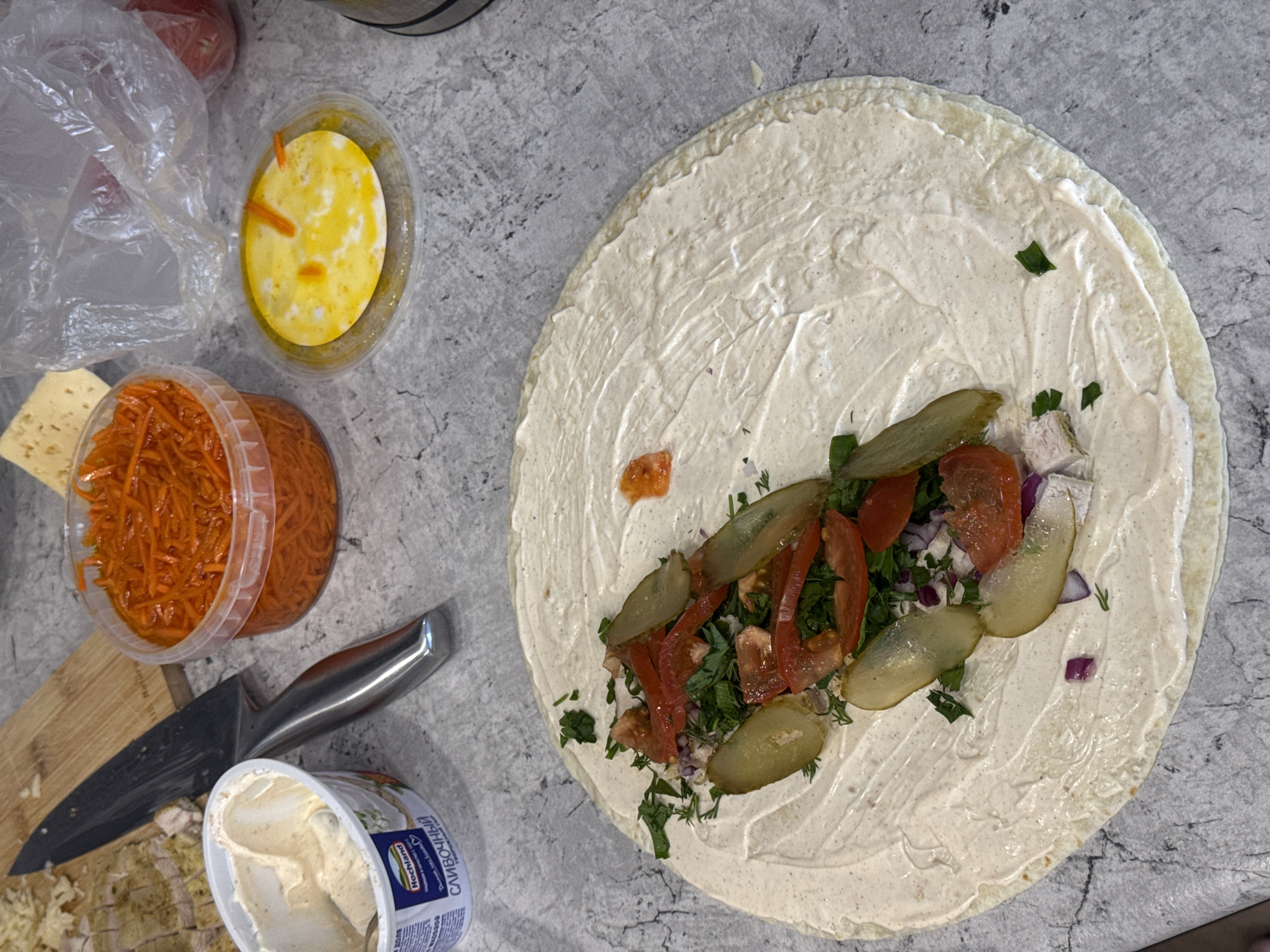 The image shows a countertop with a large tortilla spread with a white sauce. The tortilla is topped with sliced tomatoes, pickles, and parsley. A container of shredded carrots, a container of sliced cheese, a knife, and a container of white sauce are also visible. The location is unknown.