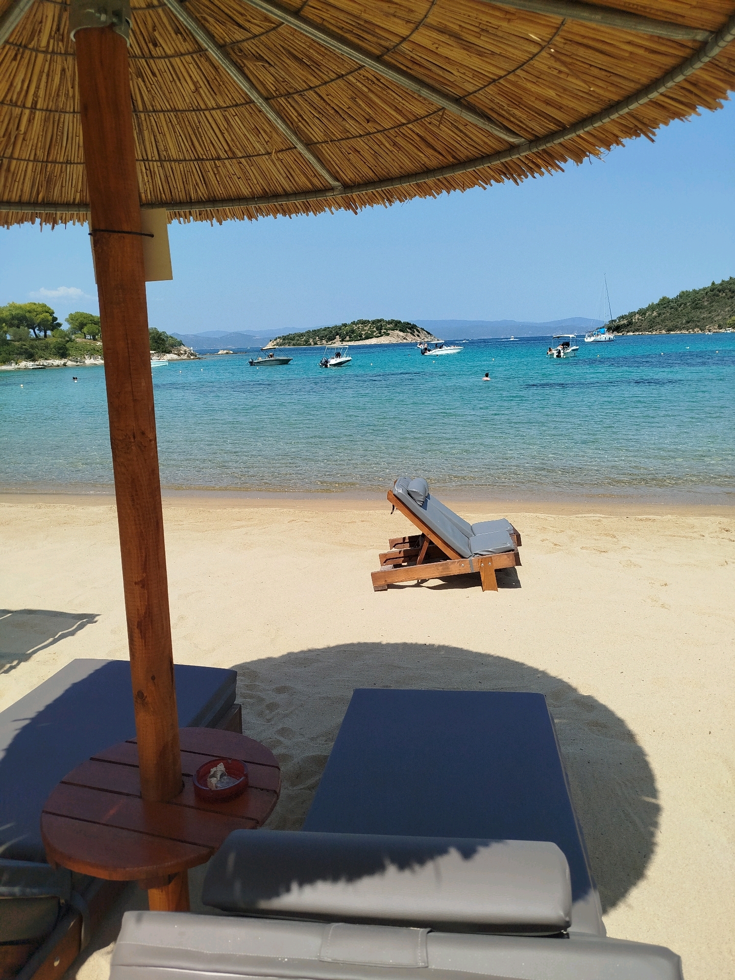 This image captures a serene beach scene in Sithonia Municipal Unit, Greece, under a clear blue sky. A large, thatched umbrella dominates the foreground, casting a dappled shadow on the light sand. Beneath the umbrella, two dark-colored lounge chairs are visible, with a small, wooden side table between them. The table features an ashtray with some cigarette butts. To the right, a single wooden and gray cushioned lounge chair is positioned closer to the water.
The turquoise water of the Aegean Sea gently laps the shore. Several boats, including speedboats and a sailboat, are dotted across the water. In the distance, lush green islands rise from the sea, creating a picturesque backdrop. The beach itself is clean and uncrowded, with only a few people visible in the water, enjoying the refreshing sea. The overall atmosphere is one of tranquility and relaxation.