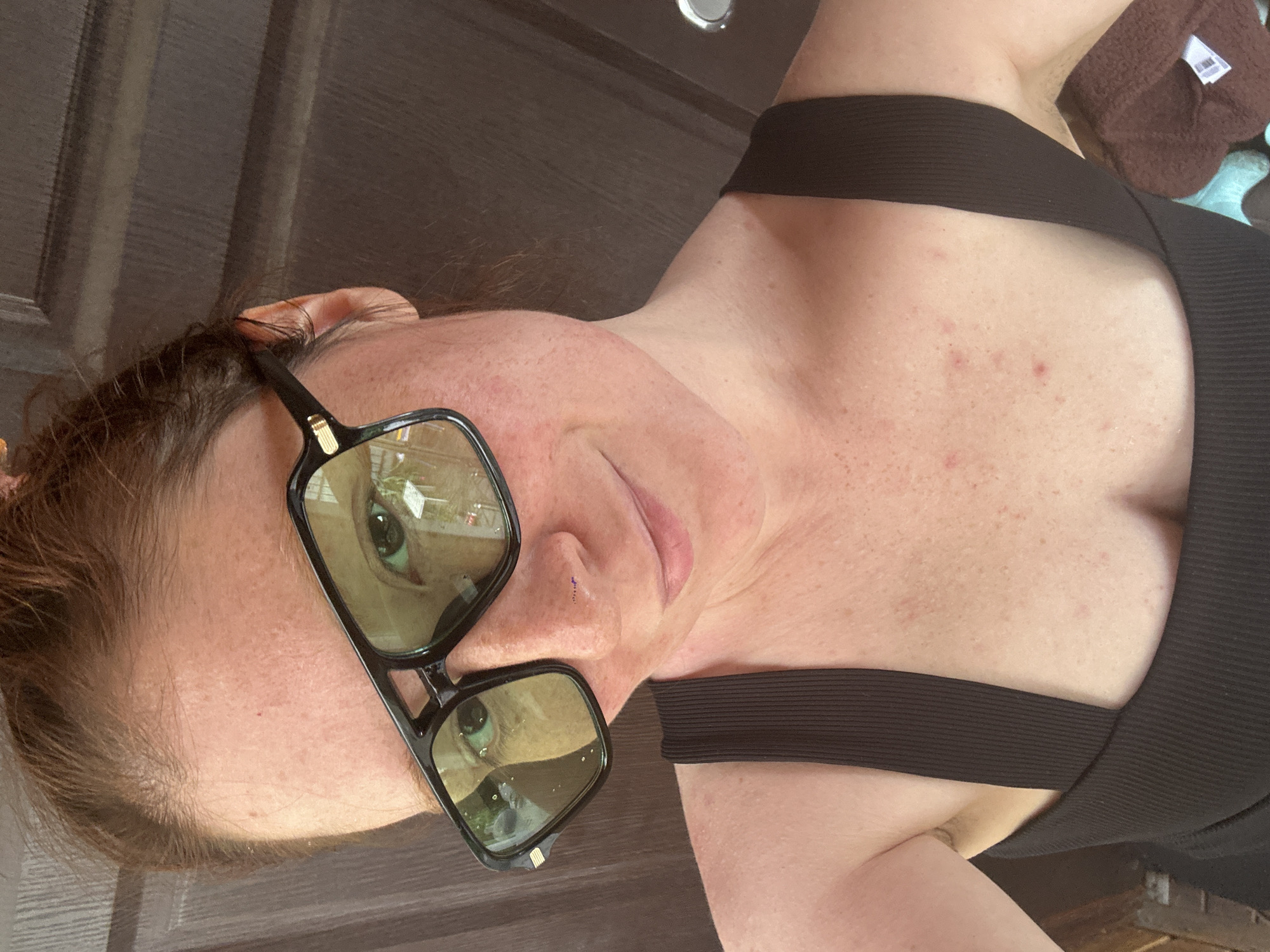 A woman is posing for a close-up photo, wearing a black tank top and large, dark-rimmed sunglasses with green-tinted lenses. Her skin is fair and freckled. She has reddish-brown hair. The reflection in her sunglasses shows an outdoor scene with greenery. The background is a dark brown, possibly a door or wall. There is no evidence of the location being Nizhny Novgorod, Russia, or any specific city.