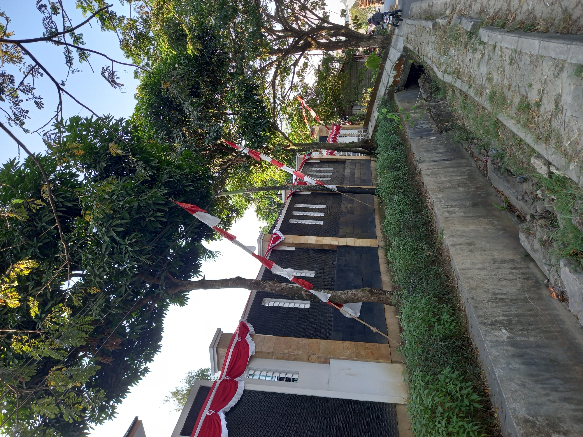 In Samarinda, Indonesia, a street scene unfolds with a concrete drain running alongside a paved walkway and road. Red and white Indonesian flags adorn a building with a textured dark facade and white accents. Trees with dense green foliage line the street, casting shadows. A person on a motorcycle is visible on the road in the distance, heading away from the viewer. The environment appears to be a dry, possibly urban or suburban area.