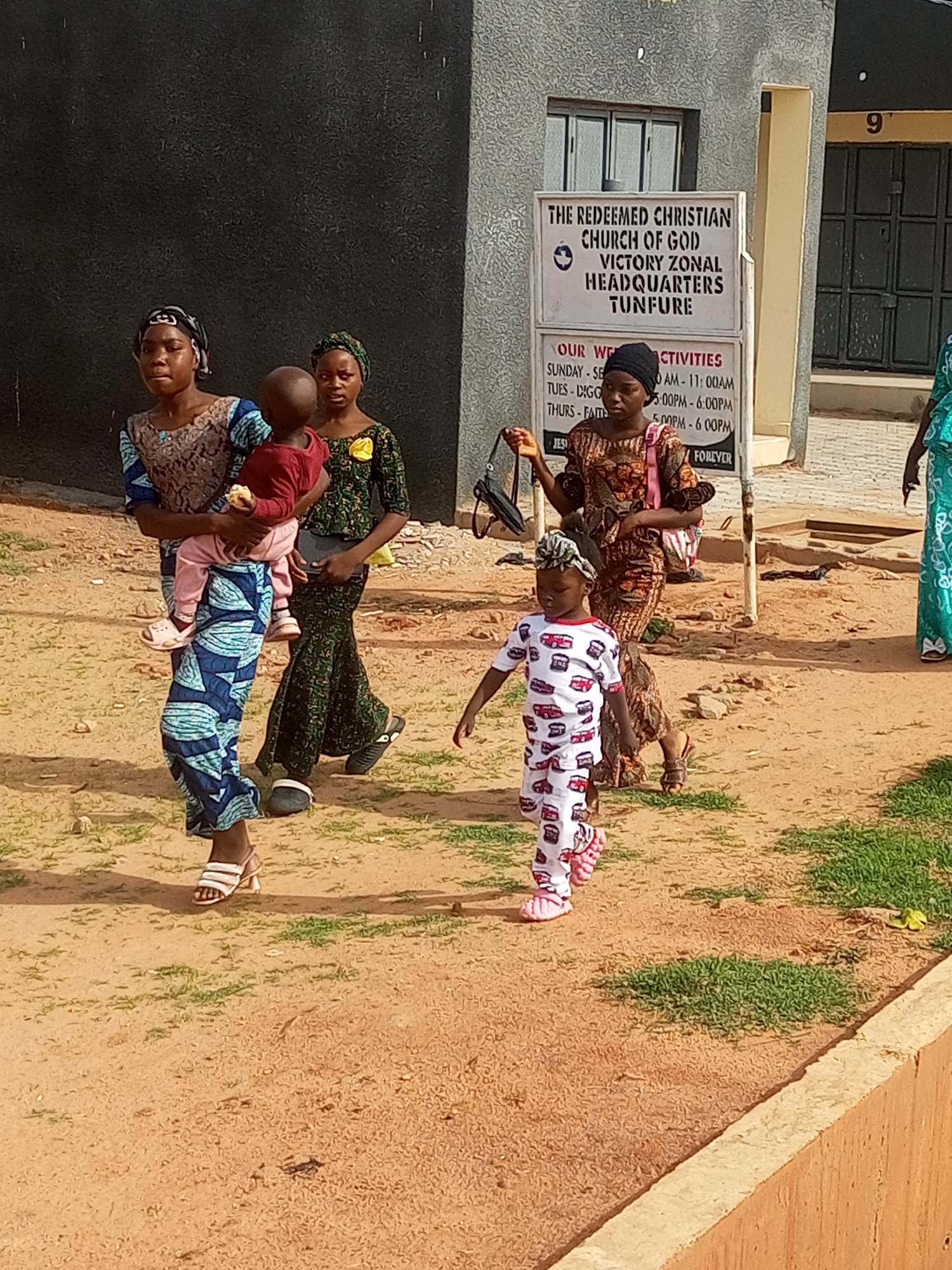 In Tempure, Nigeria, a group of individuals, likely a family, walks on a dirt pathway outside a building with a sign that reads "THE REDEEMED CHRISTIAN CHURCH OF GOD VICTORY ZONAL HEADQUARTERS TUNFURE". A woman on the left, wearing a blue and black patterned dress and headscarf, carries a baby in her arms. To her right, another woman in a green and yellow patterned dress and headscarf walks with her hand holding a dark handbag. In front of them, a young child in a white and red patterned outfit with a matching headwrap walks forward, and behind her, a woman in a brown and orange patterned dress and black headscarf, carrying a pink bag, follows. The environment is a dusty, outdoor setting with patches of dry grass.