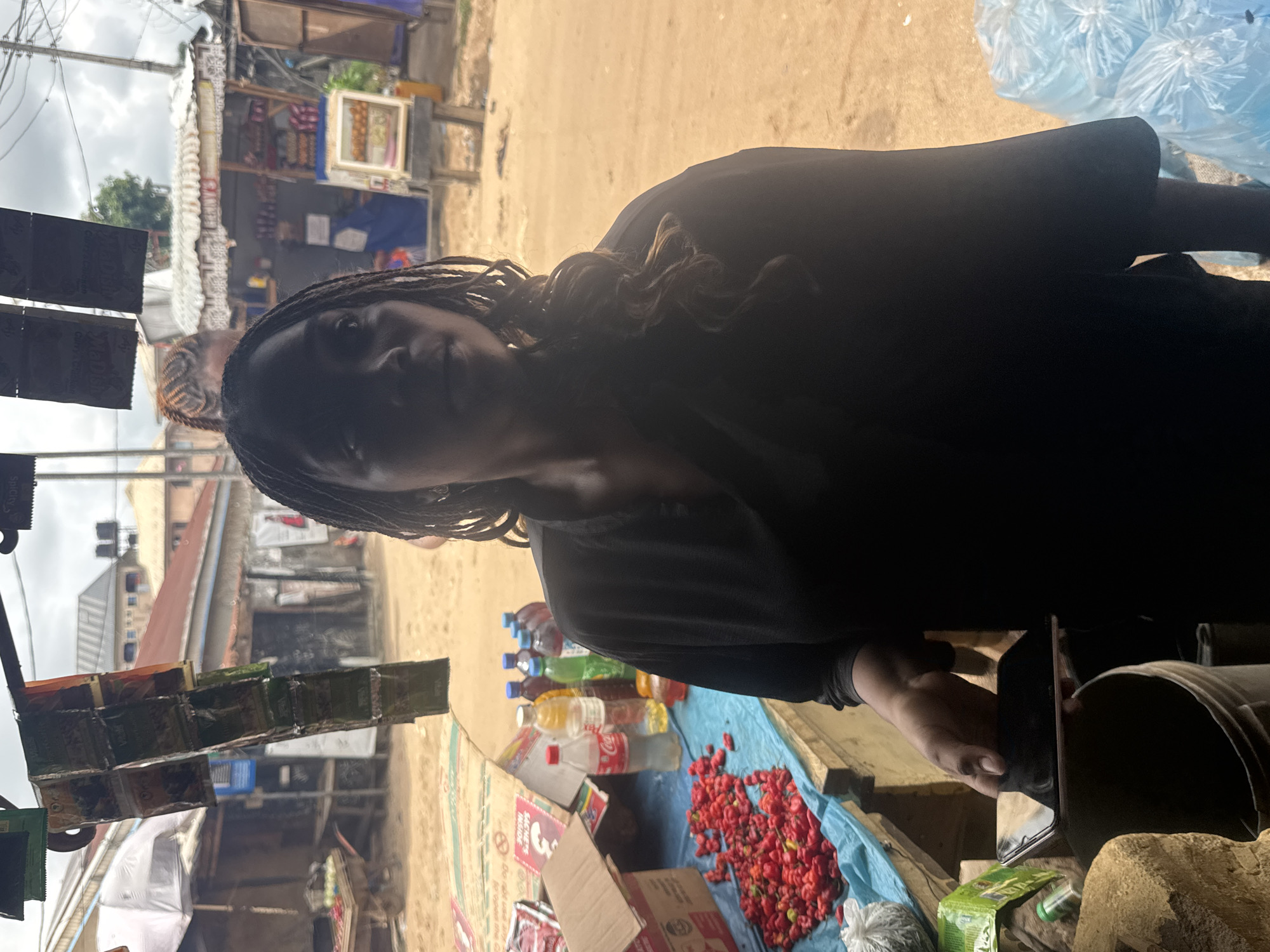 A woman stands at her roadside stand in Ihiagwa, Nigeria, offering various goods. The stand is covered with a blue tarp, on which are arrayed various wares, including red peppers and bottles of soft drinks. Packaged goods hang on display in front of the stand. The surrounding environment shows a typical street scene of the city, with buildings of various states of repair. In the background, other stands and passersby can be seen.
