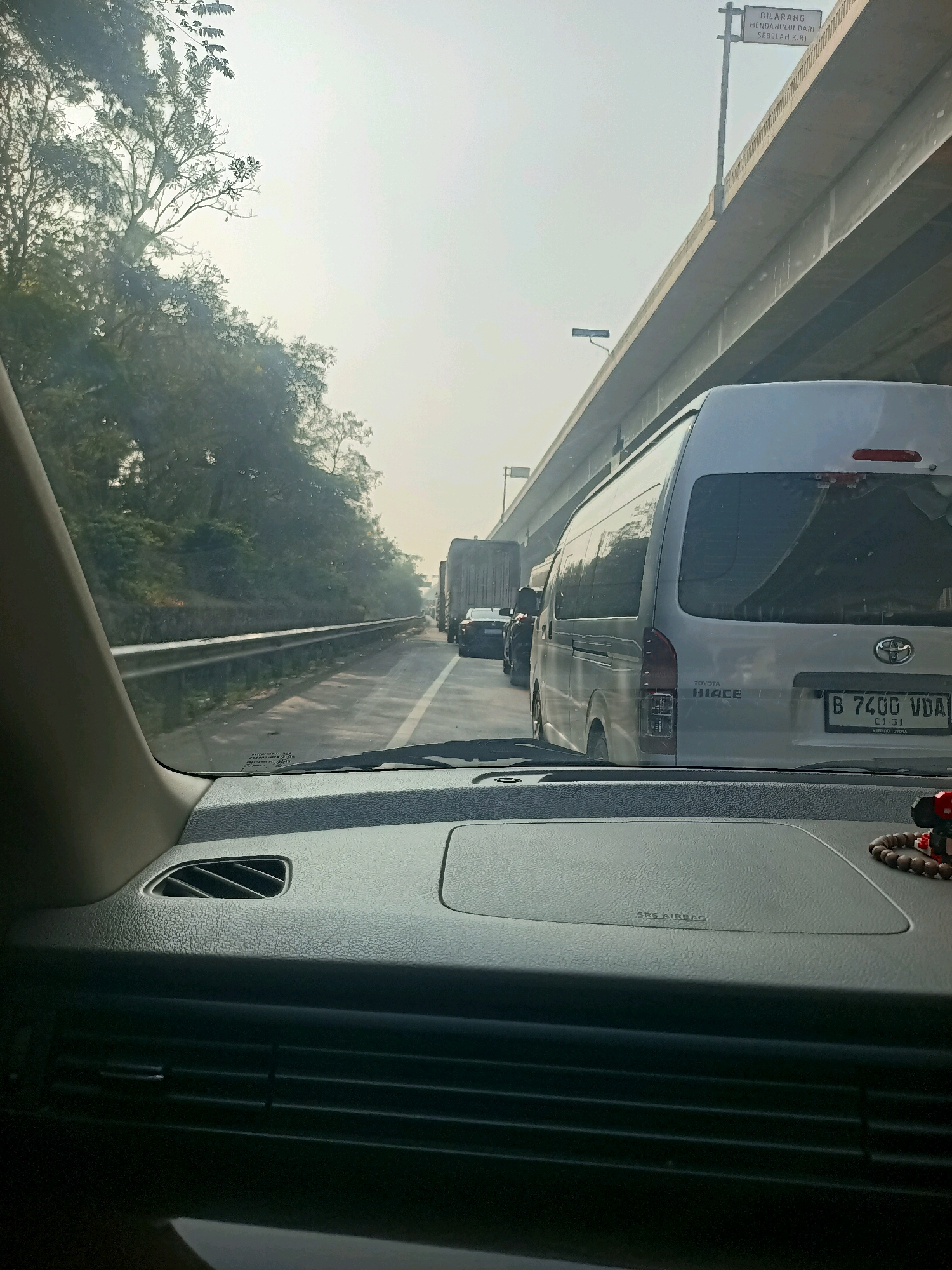 The image shows a congested highway in Cibatu, Indonesia, viewed from the interior of a vehicle. The perspective is from the driver's seat, looking forward through the windshield. The road is filled with stationary traffic, including several vehicles such as vans, trucks, and cars. A white Toyota Hiace van is prominently visible in the foreground, with its license plate reading "B 7400 VDA". The environment appears hazy, possibly due to pollution or fog, with trees and vegetation lining the left side of the road. An elevated highway structure is present on the right, with a sign on a pole that reads "DILARANG MENDAHULUI DARI SEBELAH KIRI" (Overtaking from the left is prohibited). The interior of the car shows parts of the dashboard, including air vents and an SRS AIRBAG label. A string of prayer beads is visible on the dashboard.