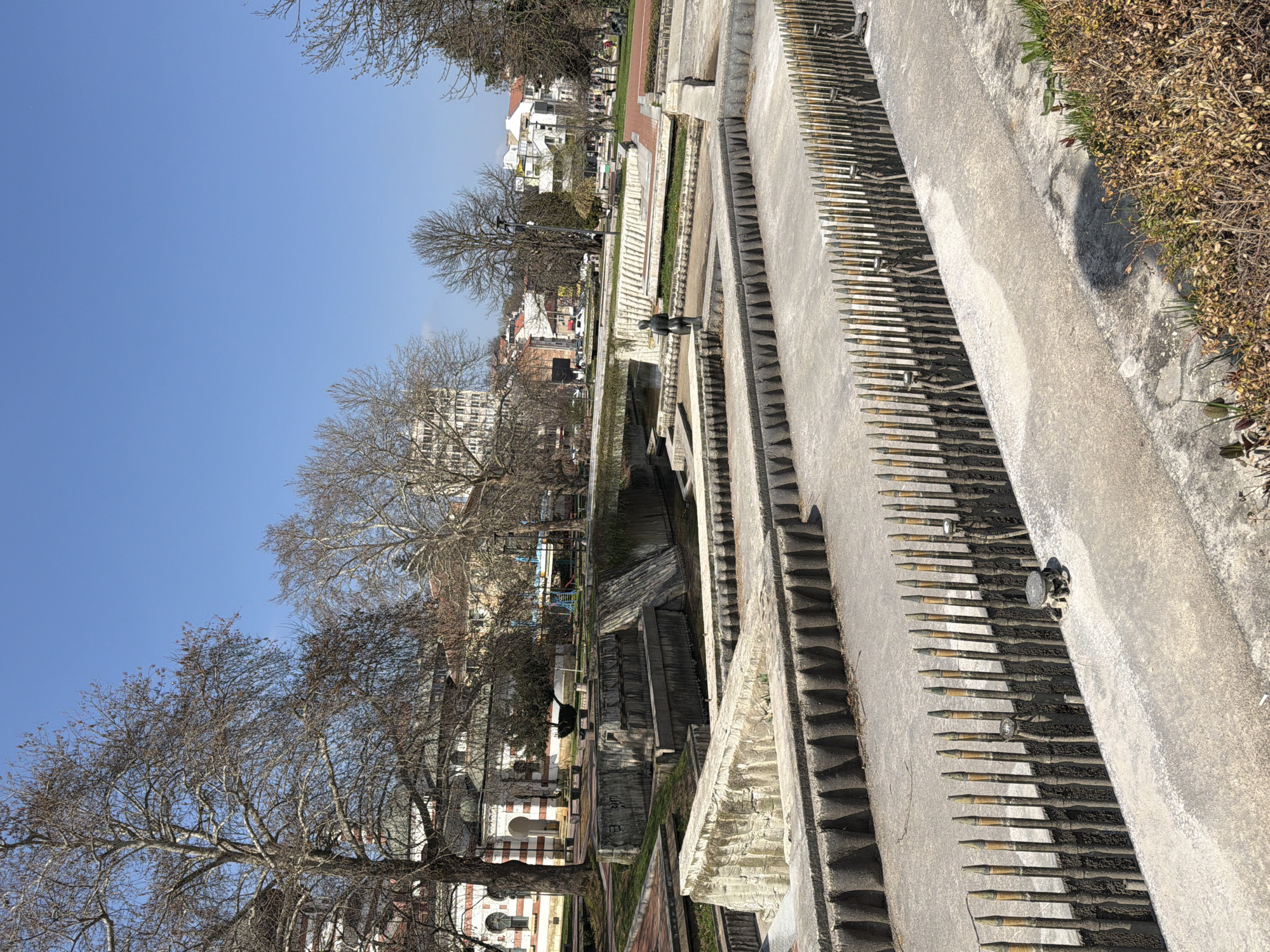 The image shows the Cascade in Pleven, Bulgaria, on a sunny day. A long, concrete structure with cascading steps and small pools forms the main subject. Some pools appear to be dry. Rows of decorative metal cylinders are affixed to the sides. In the background, buildings and bare trees are visible. A few people are walking around the site. In the foreground, a patch of dry vegetation is present. The sky is clear and blue.