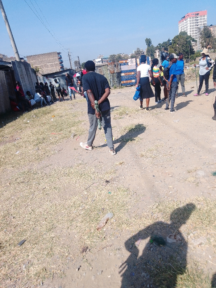 A group of people congregates in a dirt clearing amidst buildings and scattered litter. Some individuals are seated along a wall, while others are standing in smaller groups, some facing away from the camera. A person in a black shirt and gray pants holds something behind their back. The scene is bathed in sunlight, casting shadows on the ground. A tall building is visible in the background.