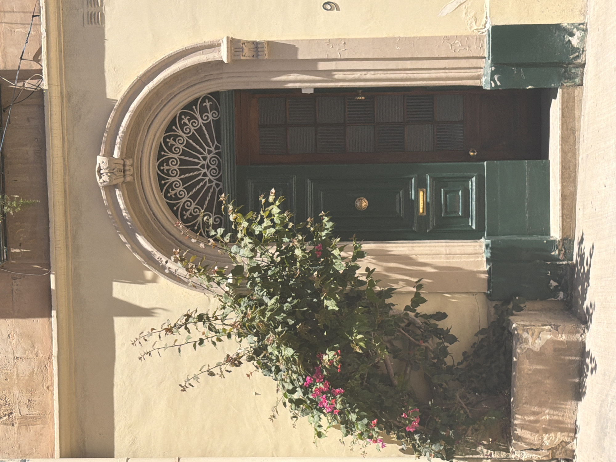 A vintage building entrance in Sliema, Malta, features a dark green, double-door entryway framed by an arched stone surround. The left door is solid and adorned with a circular brass doorknob and a brass "letters" plaque. The right door is a narrow panel with multiple small, frosted glass panes. Cascading over the entrance from the left is a vibrant bougainvillea plant with pink and magenta flowers and abundant green foliage. The building's exterior walls are painted a pale yellow, with decorative stone elements around the arch. A stone planter box sits at the base of the wall on the left. The sunlight casts sharp shadows on the ground and the building. A house number "9" is visible on the right side of the wall. The overall scene evokes a sense of aged charm and Mediterranean character.