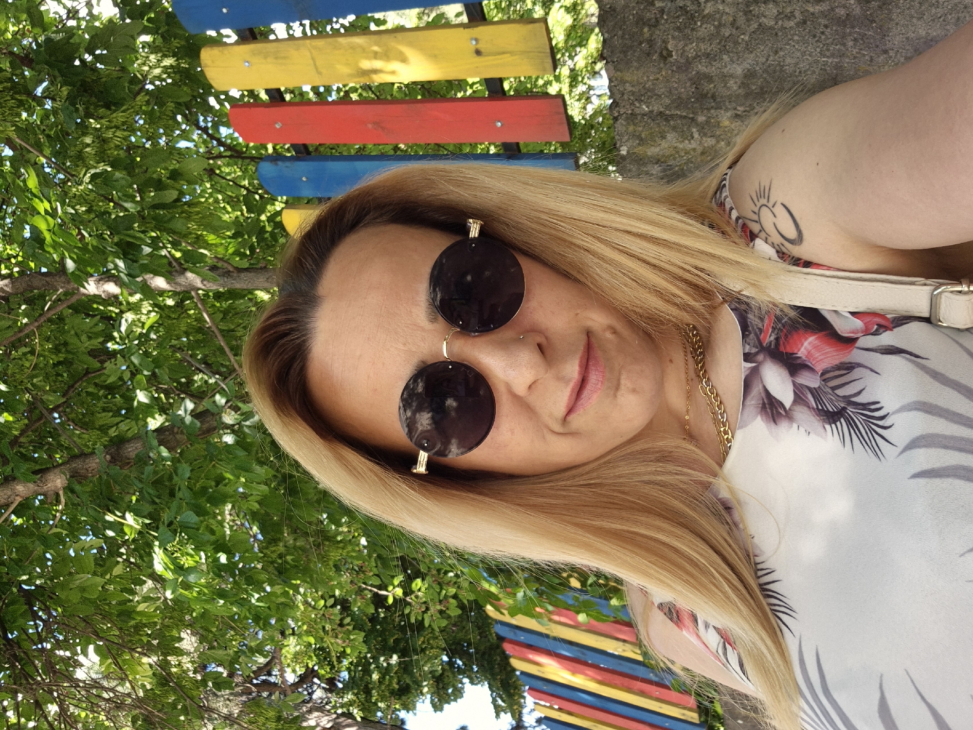 A woman with blonde hair and sunglasses is posing for a selfie. She is wearing a floral print top and a gold chain necklace. A colorful wooden fence is visible behind her, along with trees and foliage. A concrete wall is also present. The scene appears to be outdoors in a park or recreational area. The location is Unknown, Unknown.